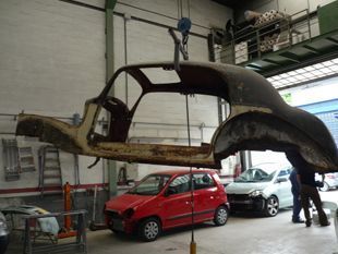 Carrocería oxidada izada en un taller sobre un coche rojo y una furgoneta blanca.