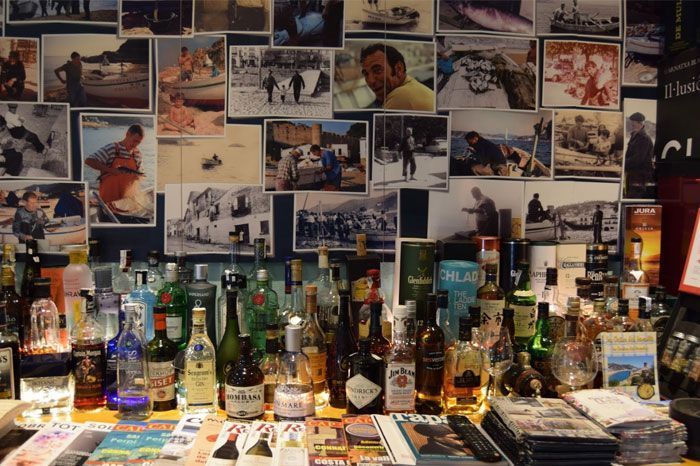 Un montón de botellas de alcohol están sobre una mesa delante de una pared de cuadros.