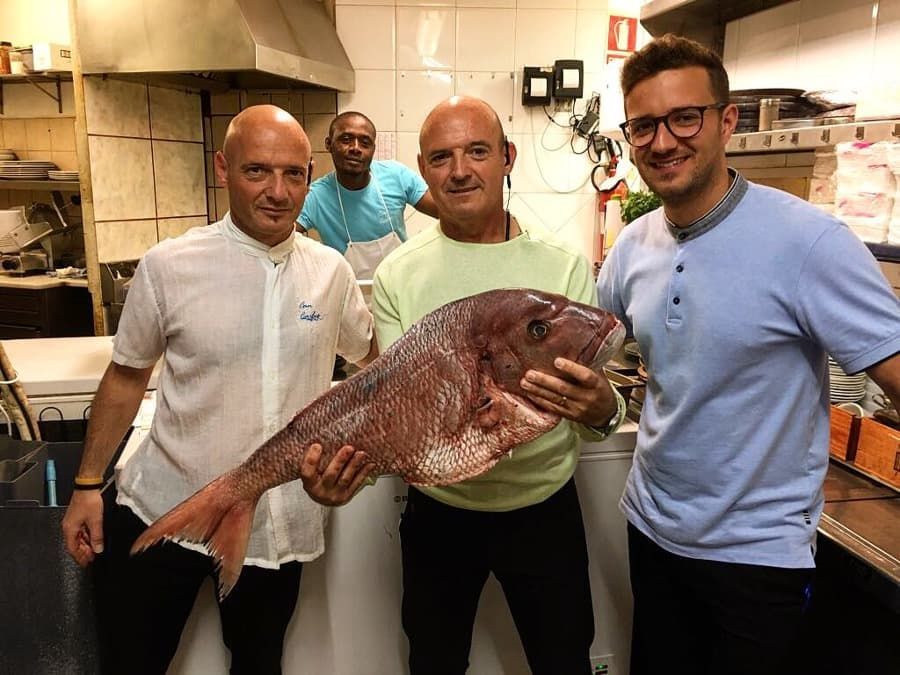 Tres hombres están parados uno al lado del otro en una cocina sosteniendo un pescado grande.