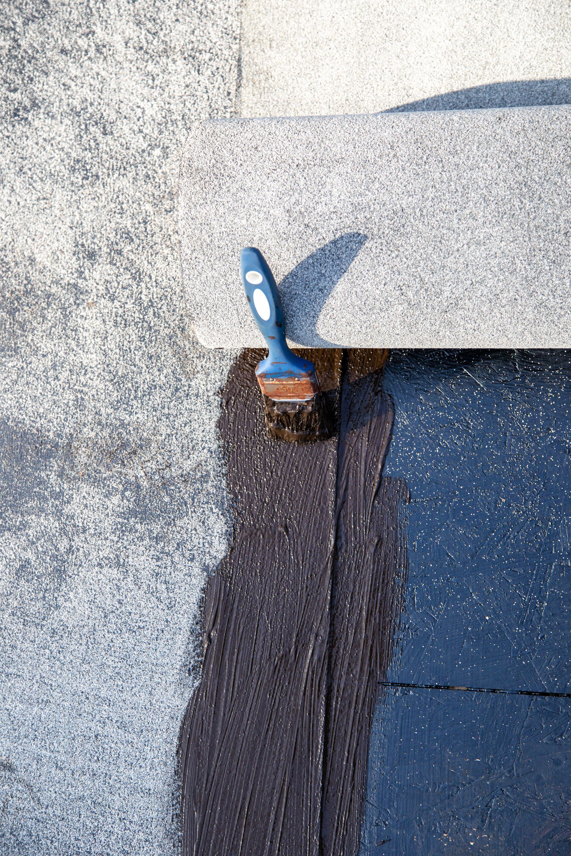 Un pinceau applique un produit d'étanchéité foncé le long du bord d'un toit plat, à l'endroit où deux couches se rejoignent.