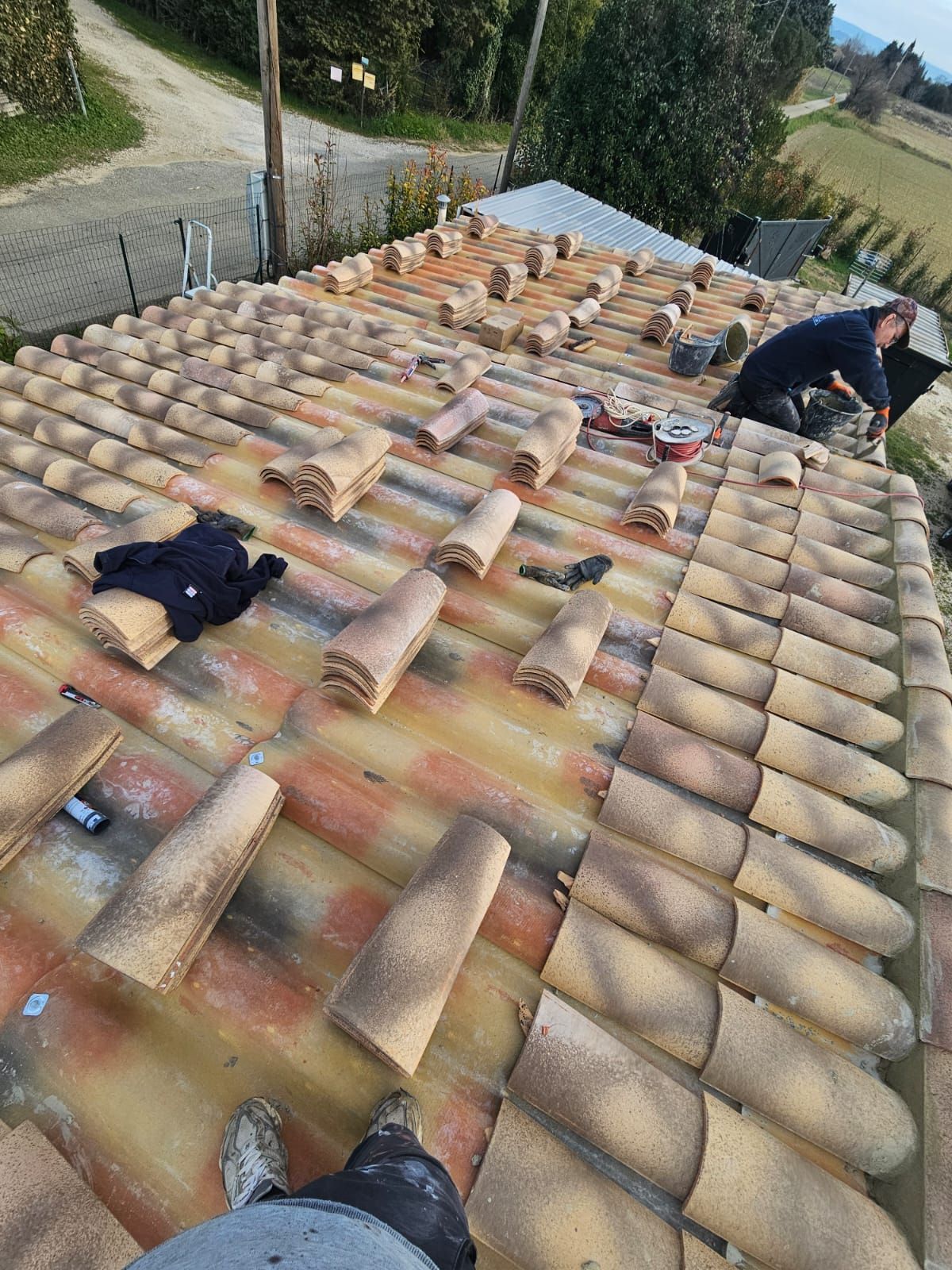 Un ouvrier pose des tuiles en terre cuite sur un toit, des piles de tuiles étant disposées de manière éparse sur la pente.