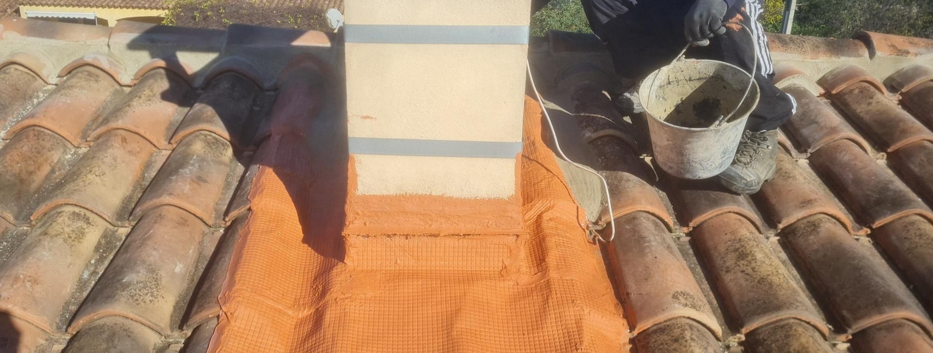 Une personne applique du mastic autour d'une cheminée sur un toit de tuiles.