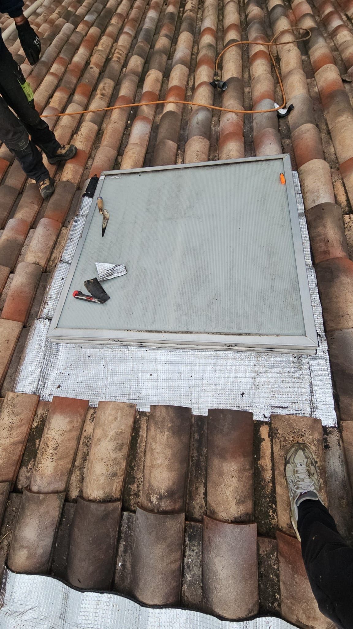 Une lucarne carrée est en cours d'installation sur un toit de tuiles en terre cuite.