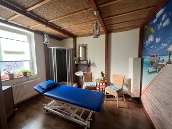 Massageraum mit blauer Behandlungsliege, Korbstühlen, Fenster und Strandmotiv unter einer Strohdecke