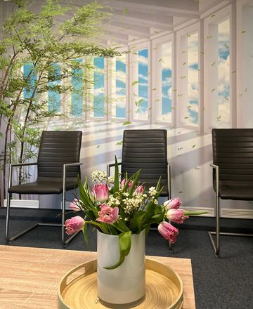 Ein Blumenarrangement auf einem goldenen Tisch in einer hellen Lobby mit schwarzen Stühlen und großen Fenstern.