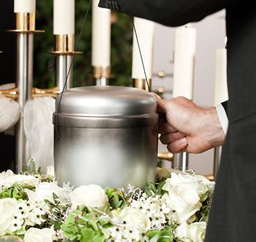 Un hombre con traje sostiene una urna de metal delante de velas y flores.