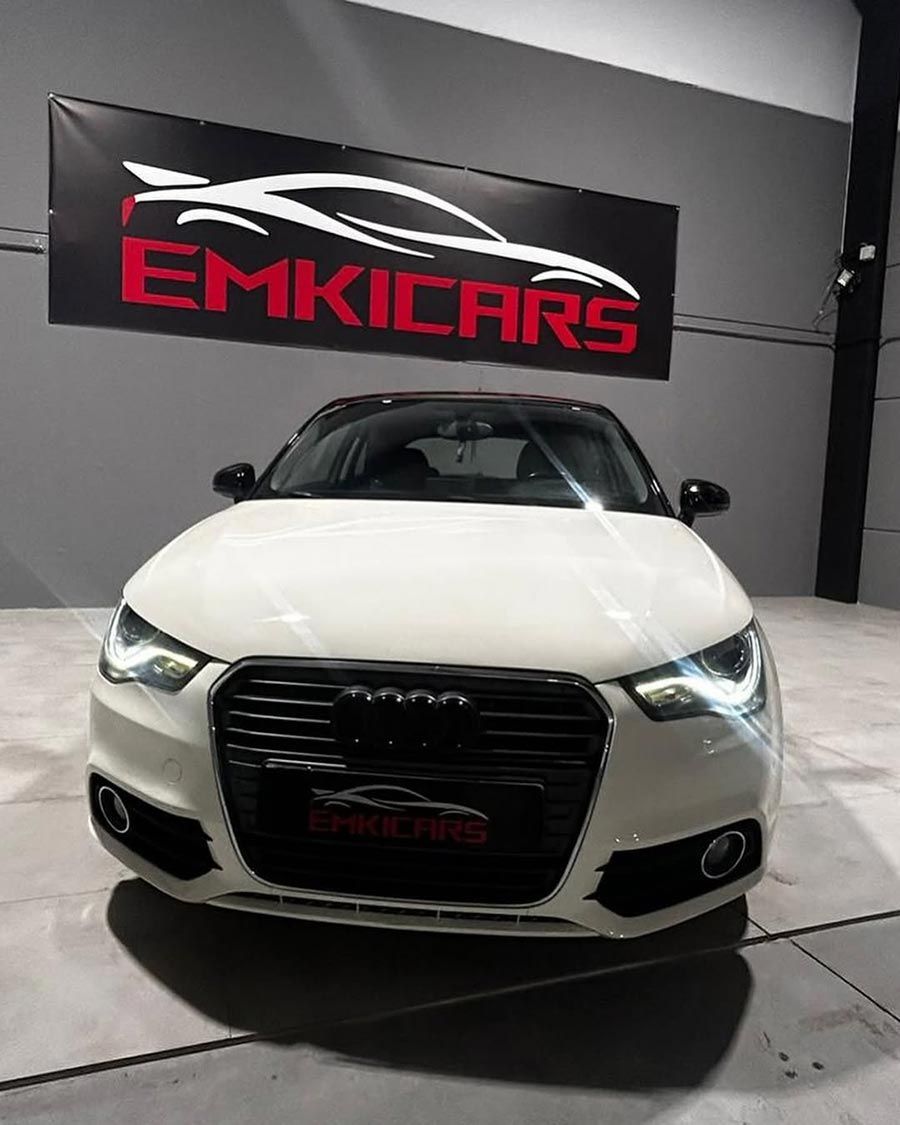 Vista frontal de un coche blanco en una sala de exposición con un letrero de EMKICARS en la pared.