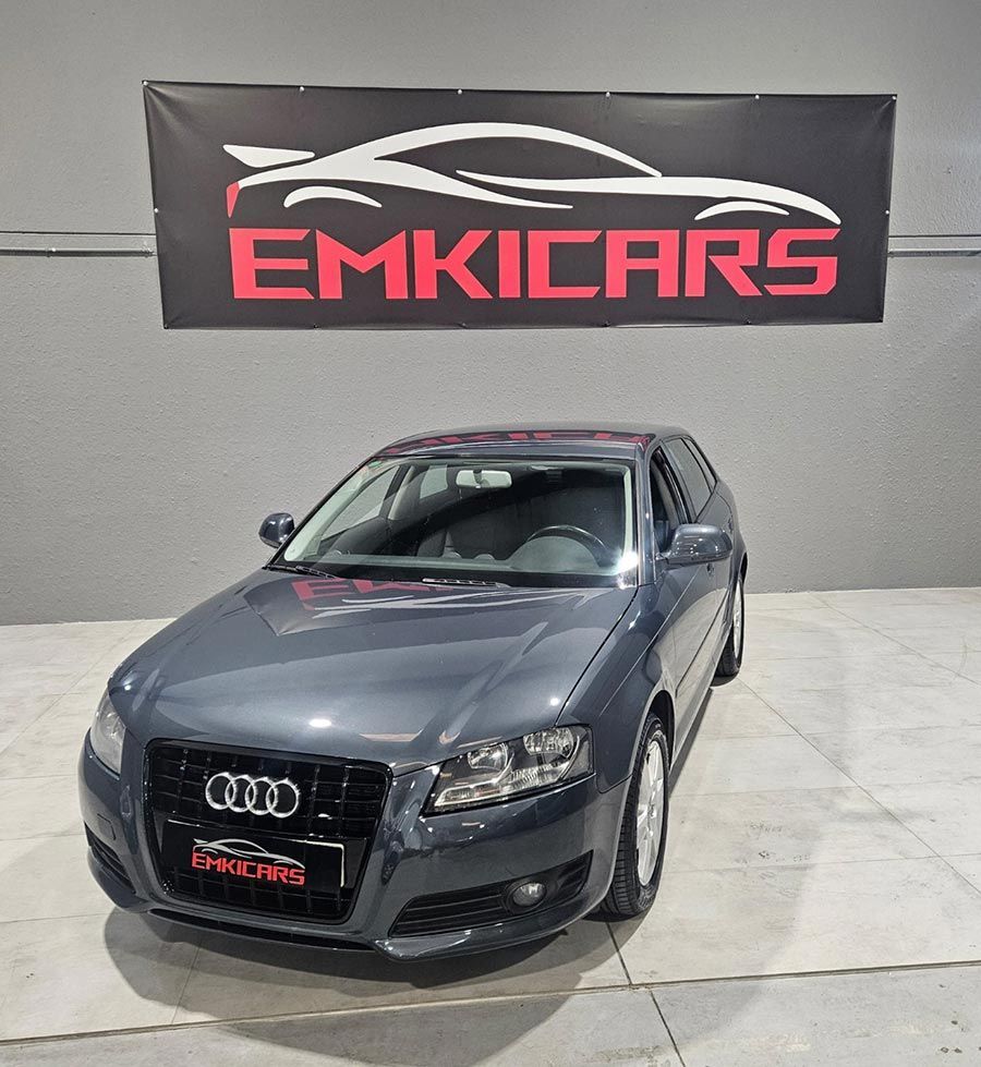 Un sedán Audi de color gris oscuro estacionado en el interior, frente a una pancarta de EMKICARS.