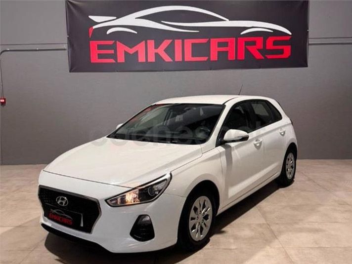 Un Hyundai hatchback blanco se exhibe en una sala de exposición con un letrero de EMKICARS en la pared.