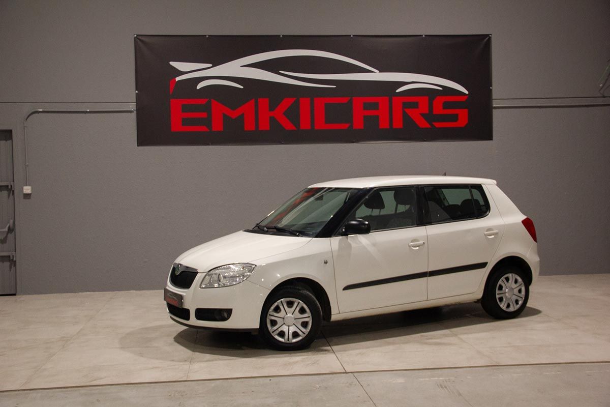 Coche hatchback blanco expuesto en una sala de exposición con un letrero de EMKICARS en la pared.