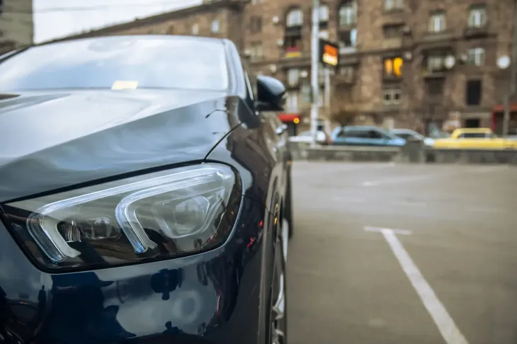 Primer plano de un faro de coche azul oscuro en un aparcamiento urbano, con edificios al fondo.