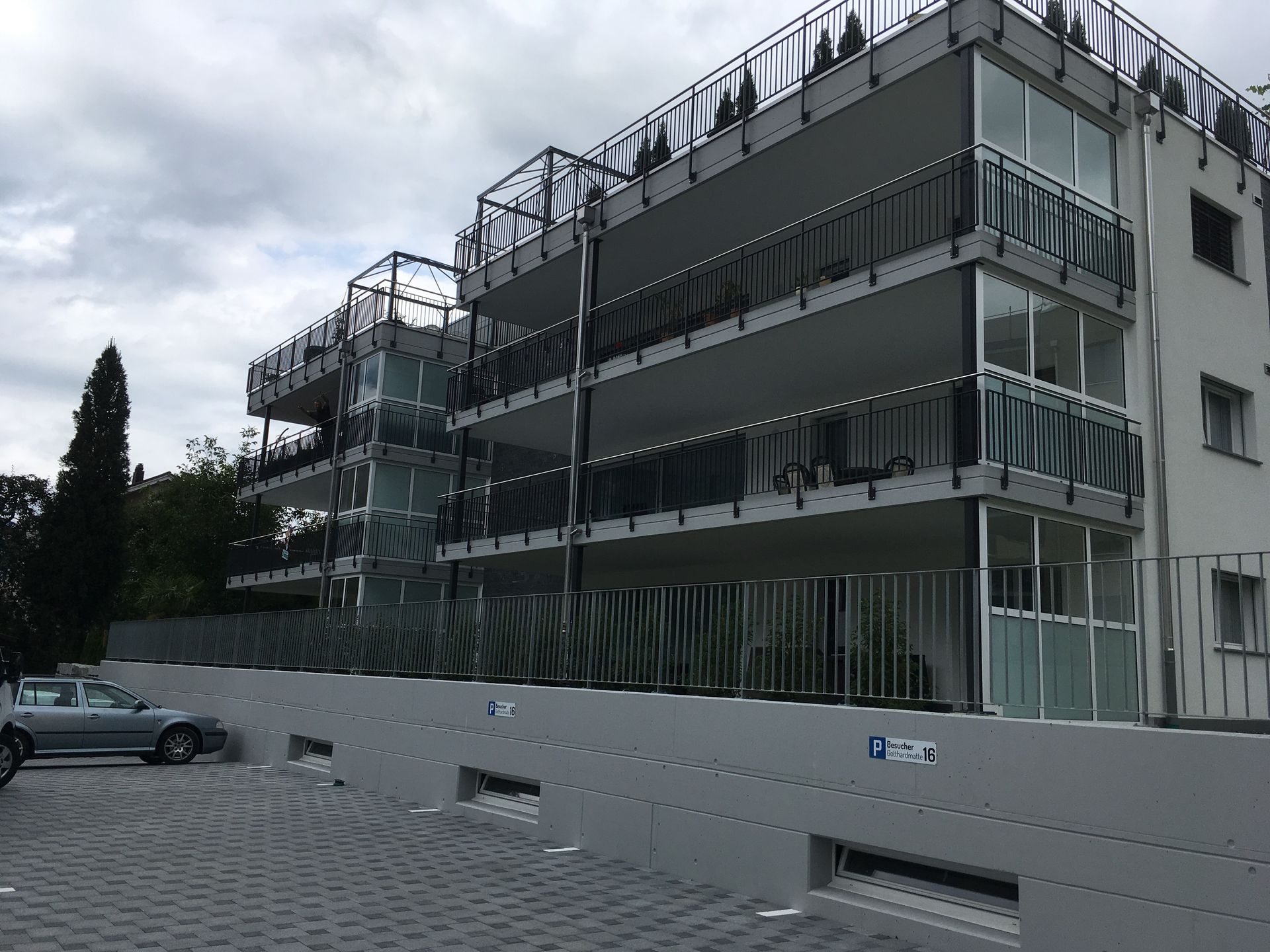 Modernes Mehrfamilienhaus mit Balkonen und Dachgeländer hinter einem geparkten Auto und einer weissen Wand