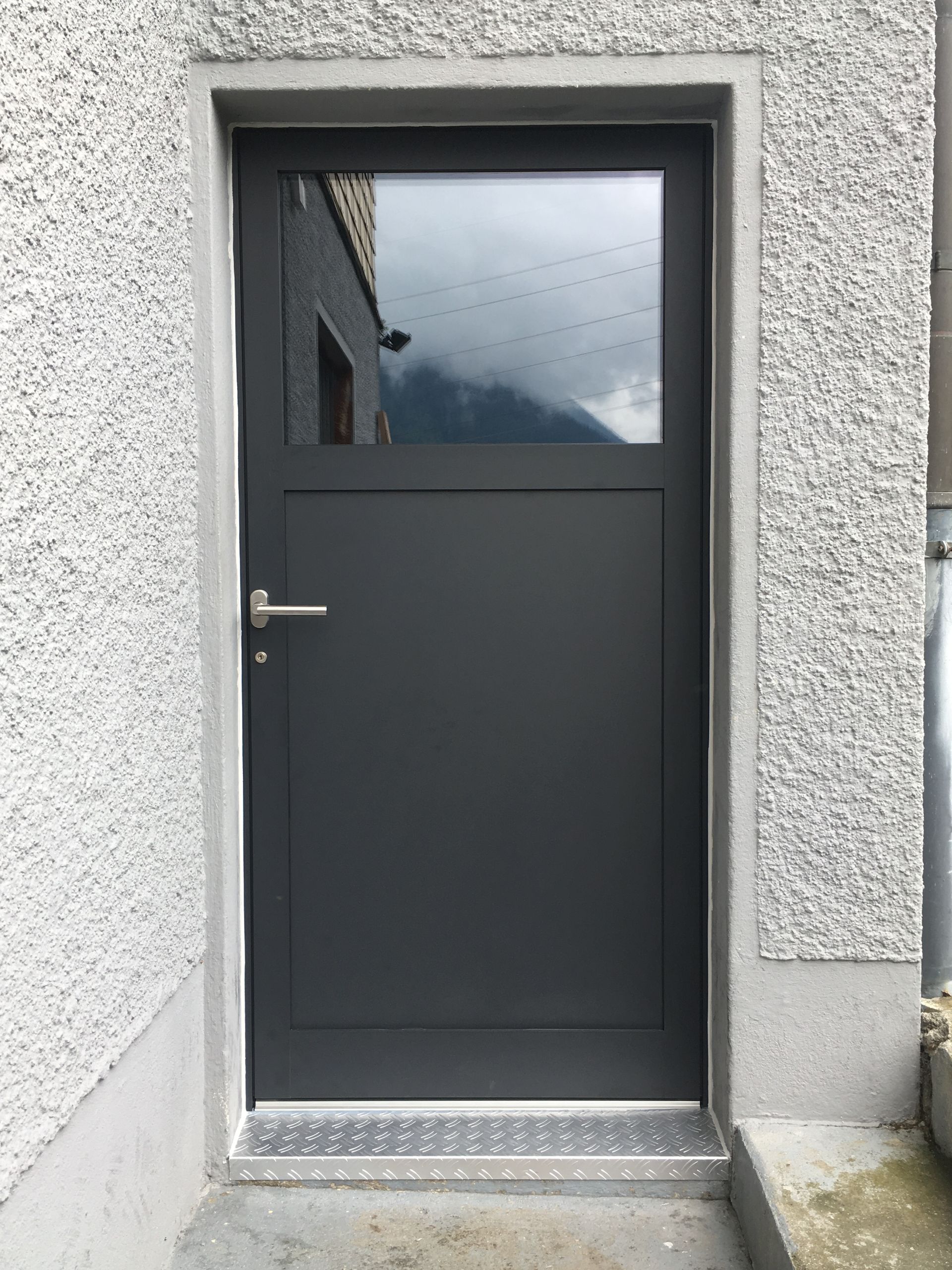 Dunkelgraue Aussentür mit Fenster, eingefasst in eine strukturierte weisse Wand und eine Betontreppe.
