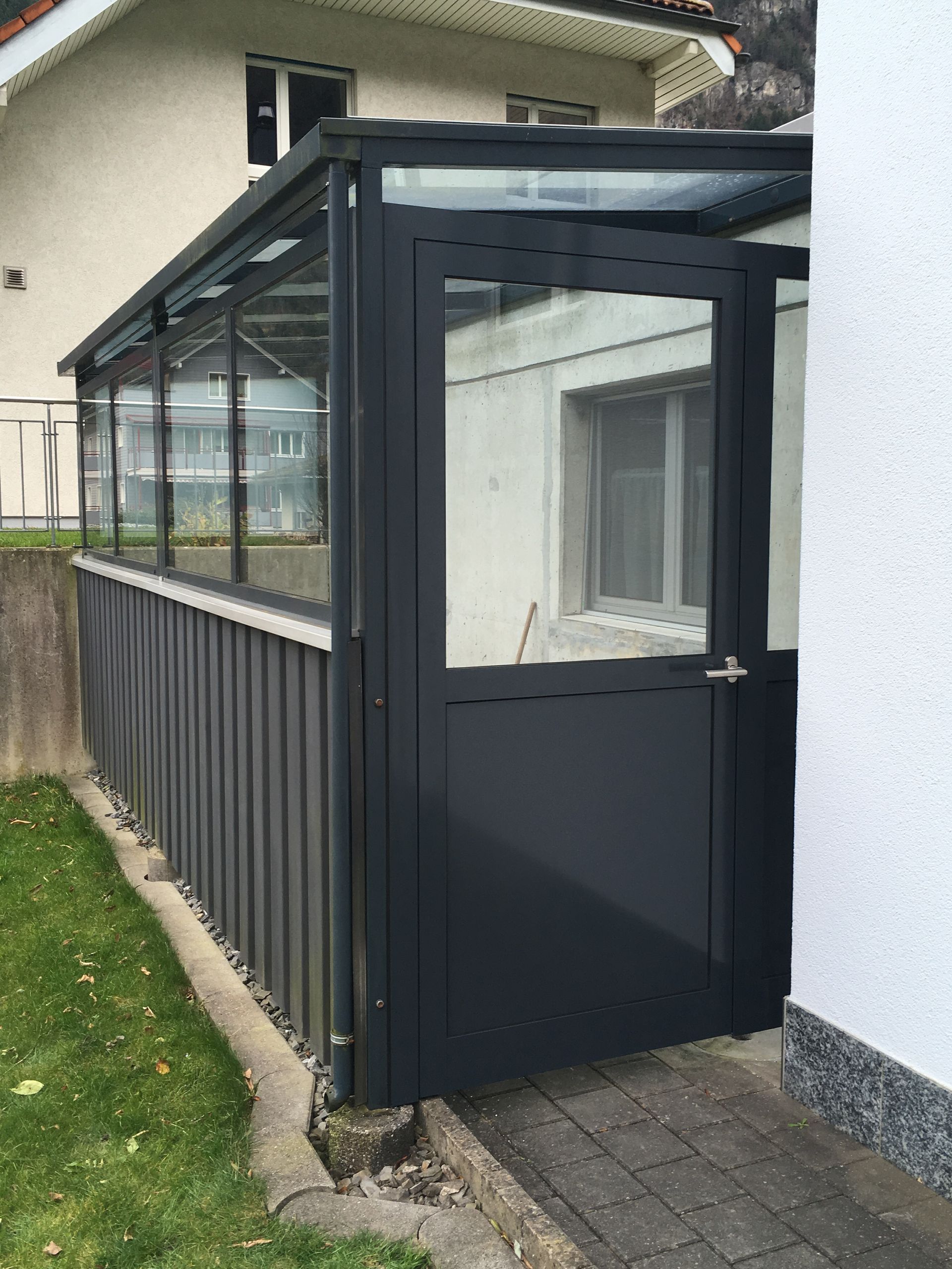 Schwarzer, geschlossener Vorraum oder Windfang, der an ein weisses Haus angebaut ist, mit Glasfenstern und einer Tür.
