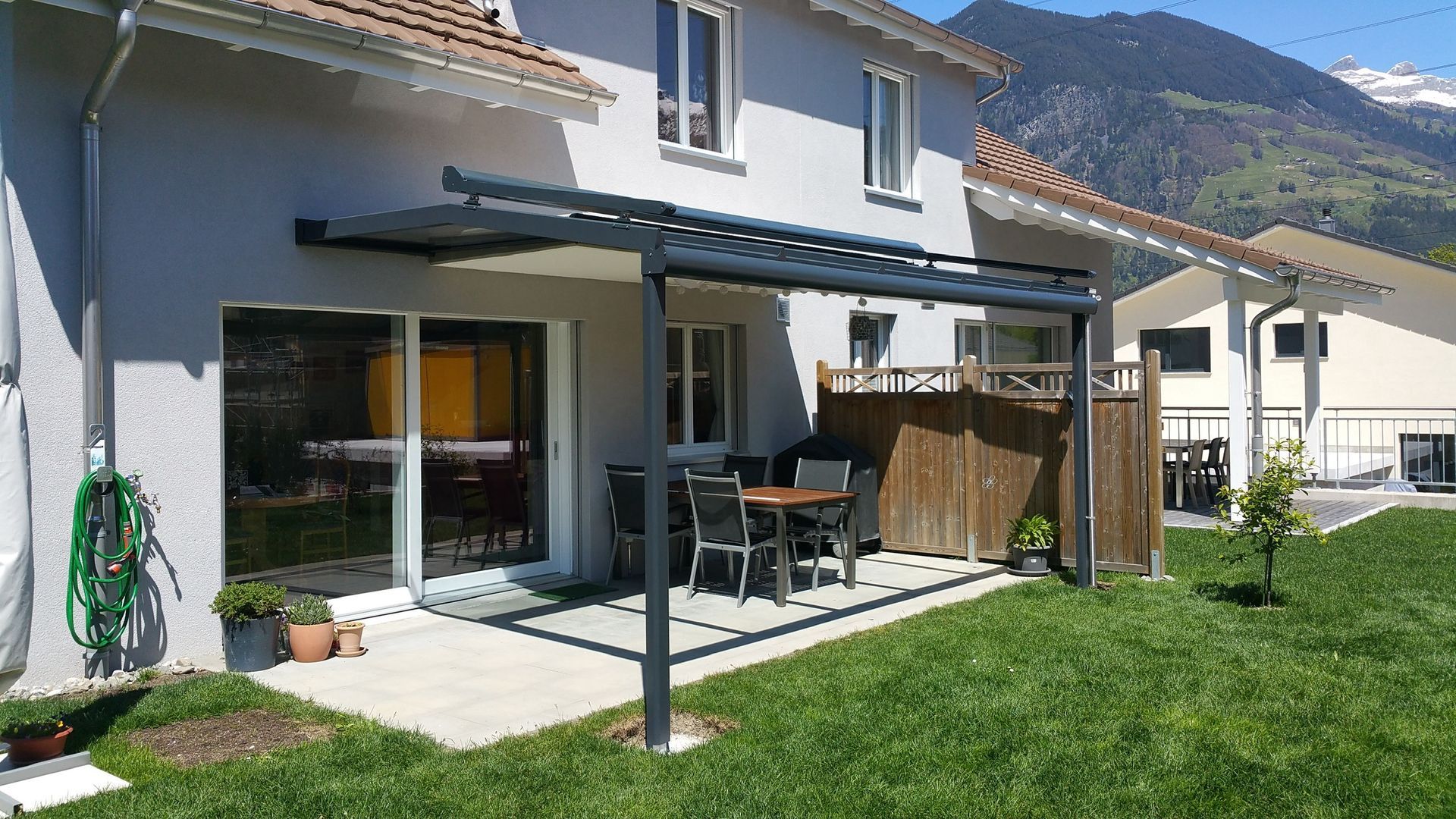 Terrasse mit schwarzer Pergola, Glasschiebetüren, Gartentisch und grünem Rasen neben einem Haus in Berglandschaft