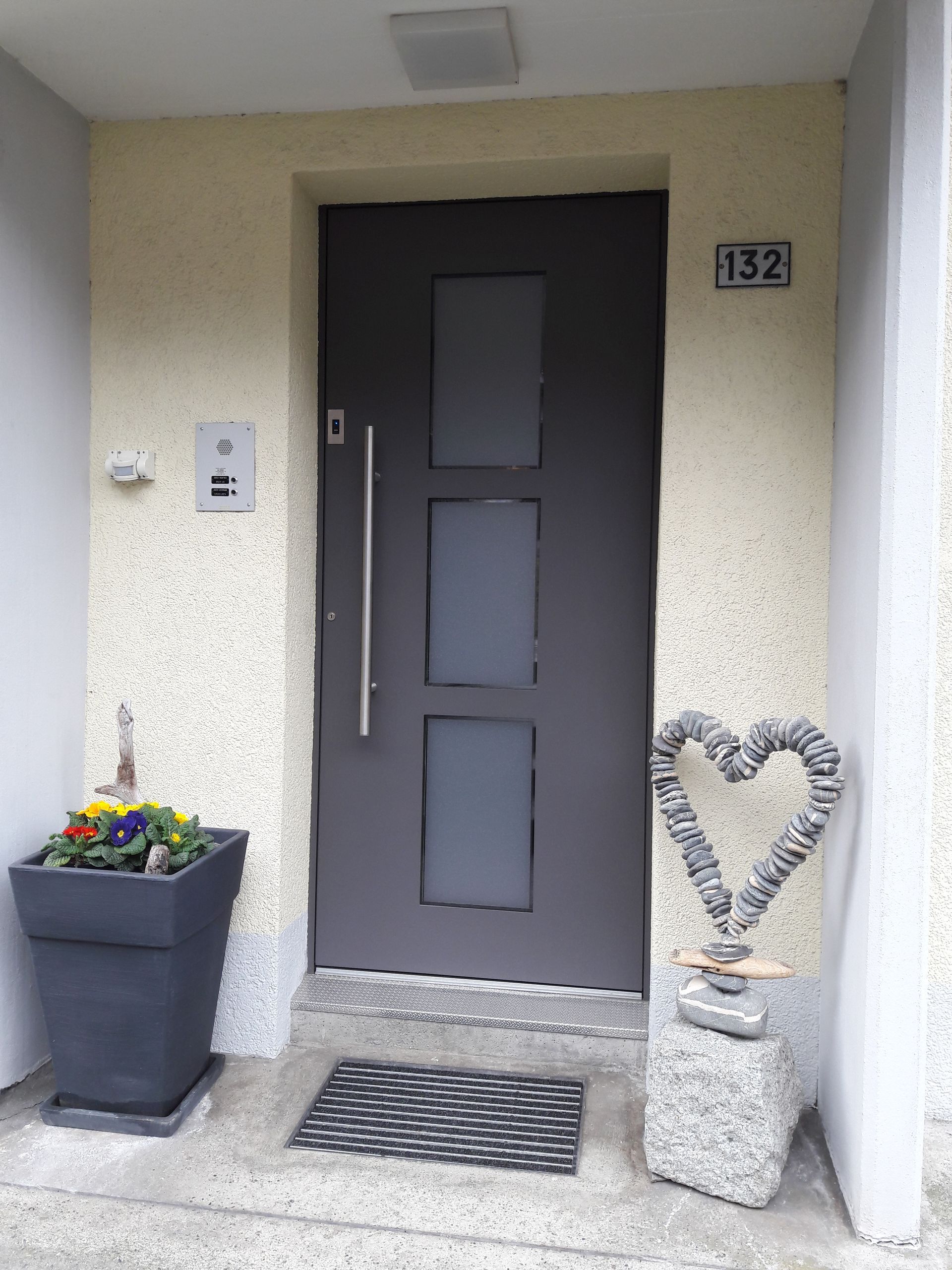 Graue Haustür mit Hausnummer 132, Topfblumen und eine herzförmige Steinverzierung am Eingang