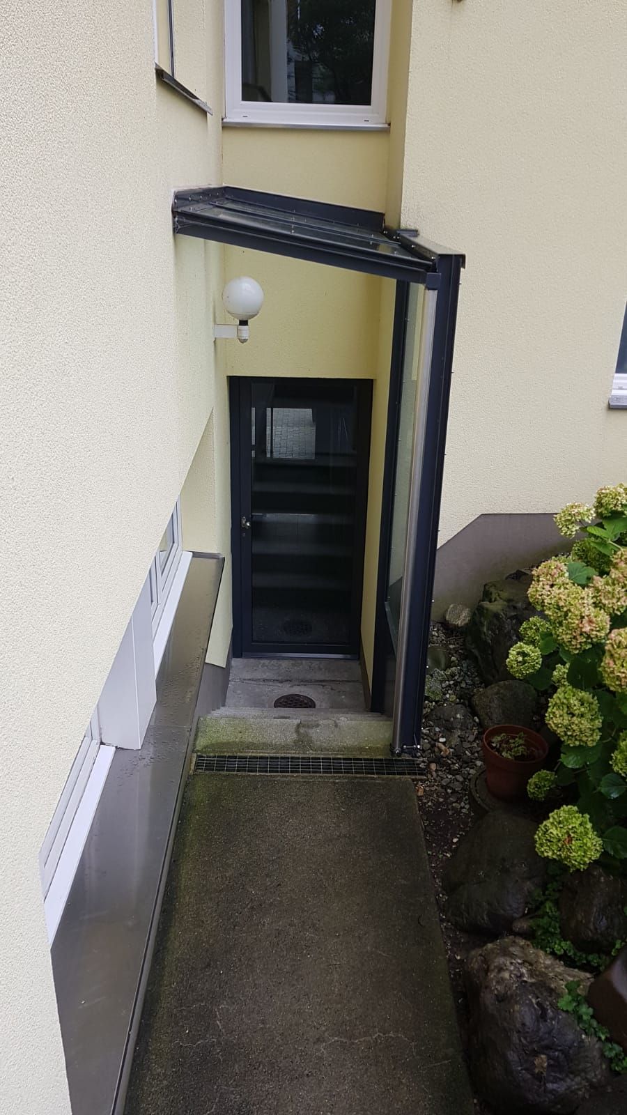Schmaler Kellereingang unter einer schwarzen Markise neben einem Haus, mit Stufen hinunter und einem nassen Betonweg.