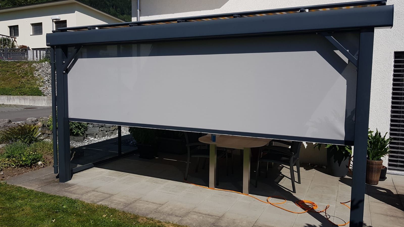 Graue, herunterrollbare Aussenmarkise überspannt eine Terrasse mit einem darunter stehenden Tisch neben einem Haus.