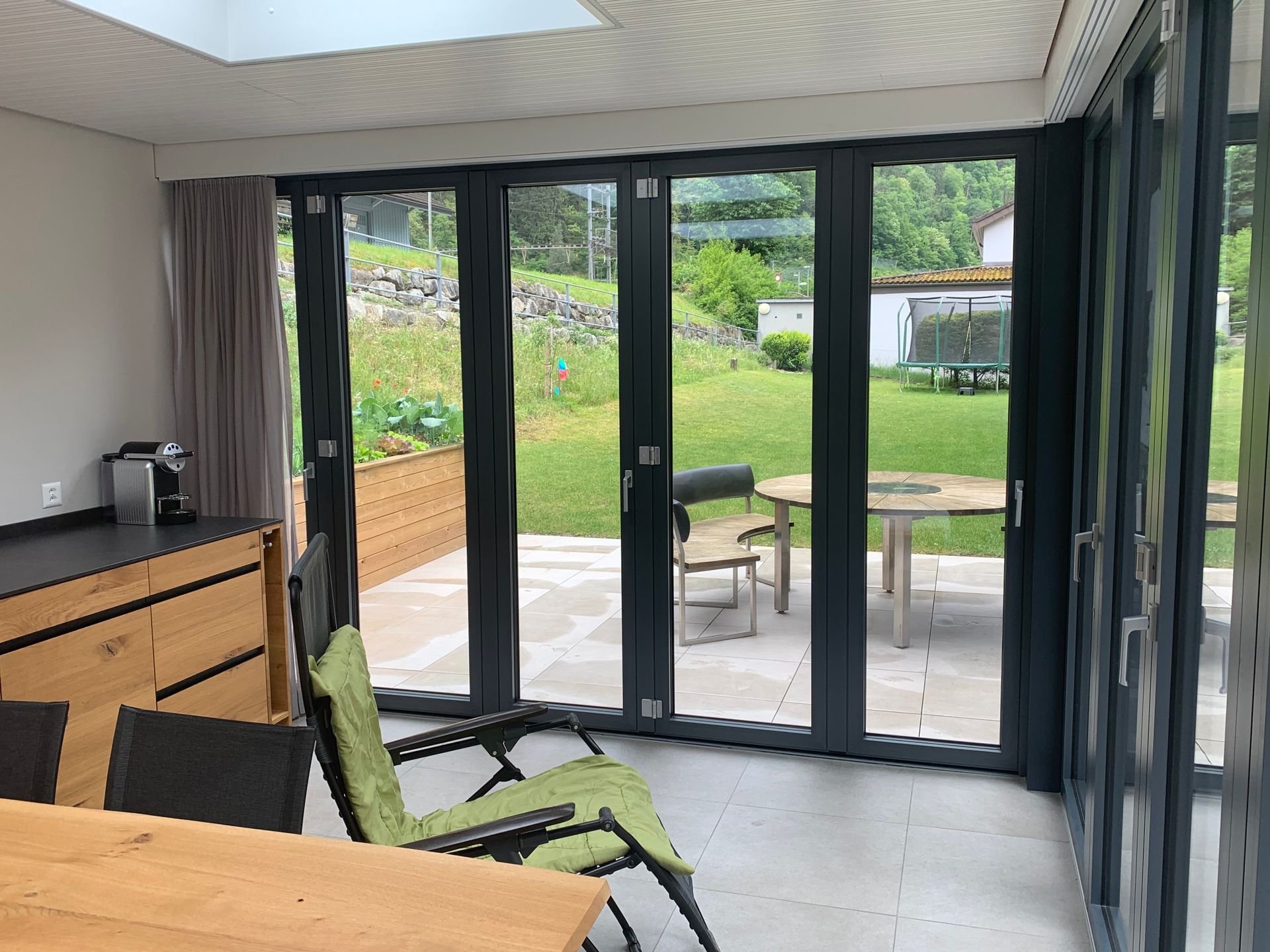Modernes Zimmer mit schwarz gerahmten Glastüren, die sich zu einer grünen Terrasse und einem Garten öffnen.