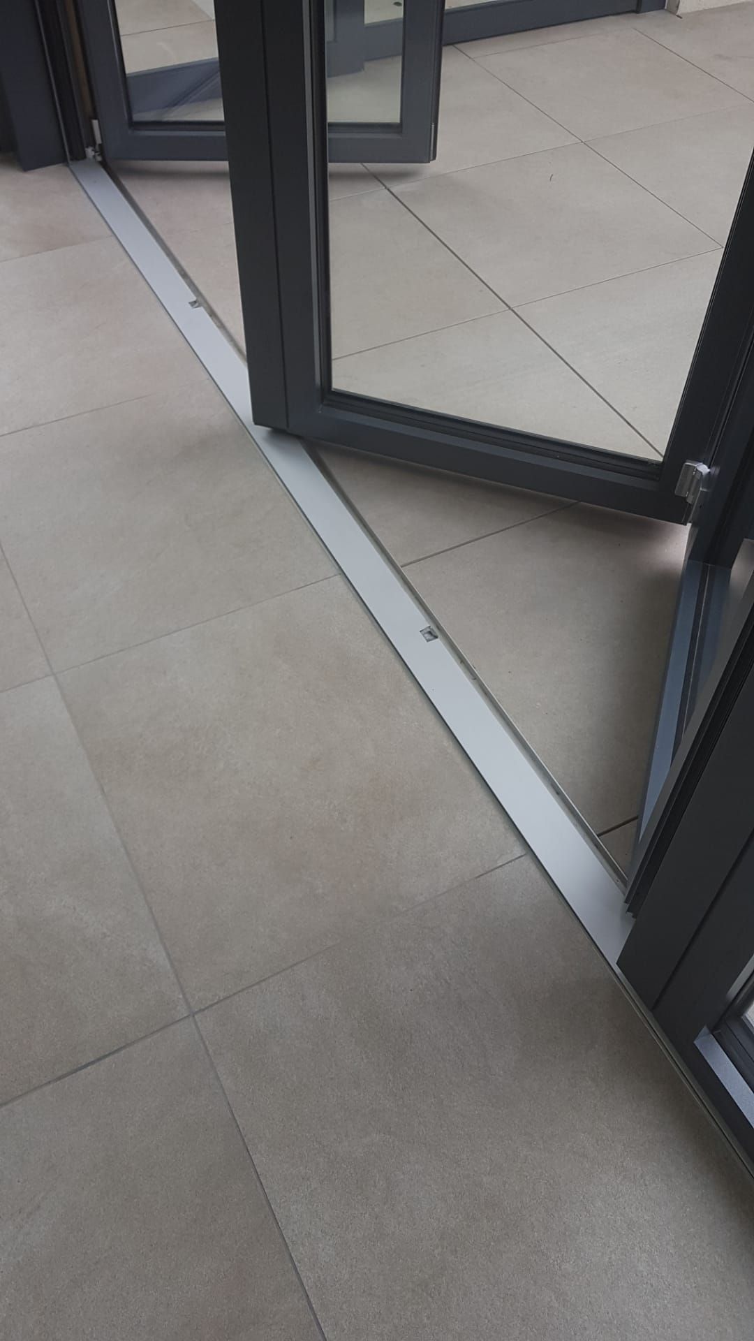 Eine Glastür öffnet sich zu einem gefliesten Boden mit einer metallischen Schwelle und einem schwarzen Türrahmen.
