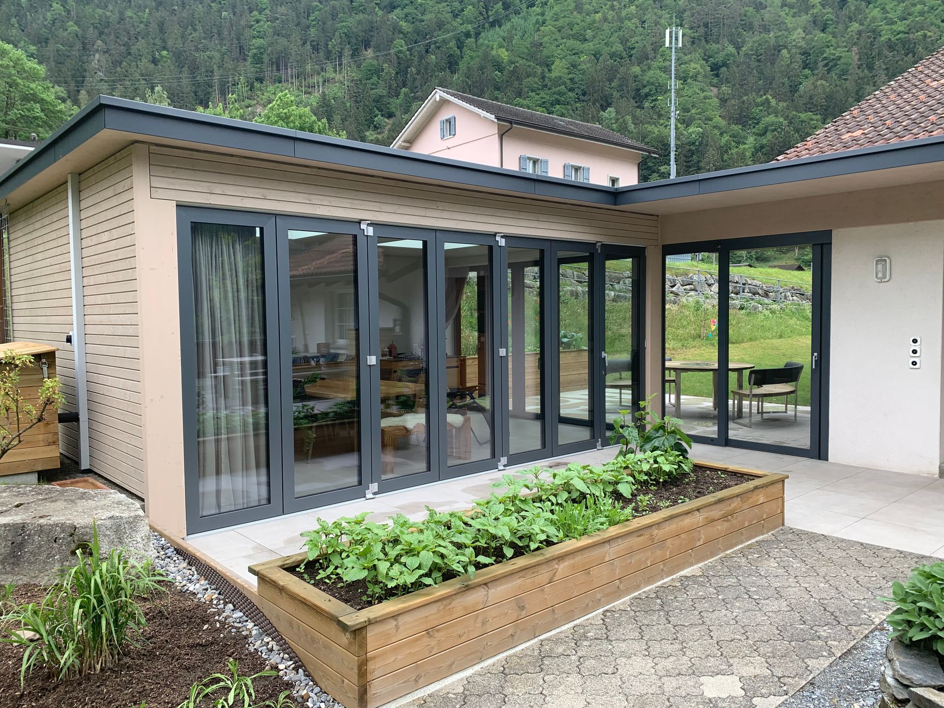 Moderner Hausanbau mit Glastüren, hölzernem Pflanzkasten und Steinterrasse mit Blick auf einen grünen Hang