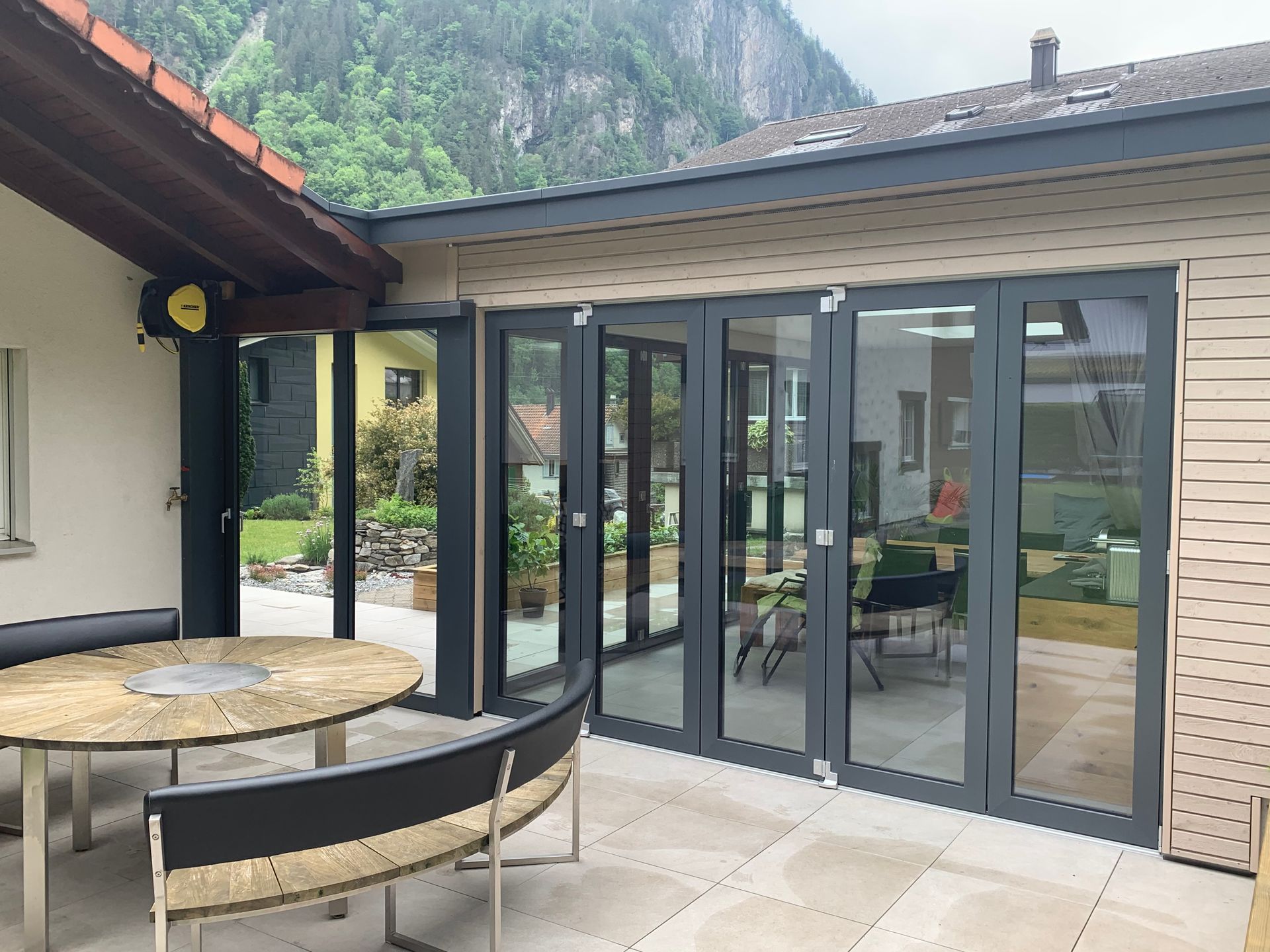 Terrasse mit rundem Steintisch, geschwungener Bank und verglastem Wintergarten neben einem Berghaus