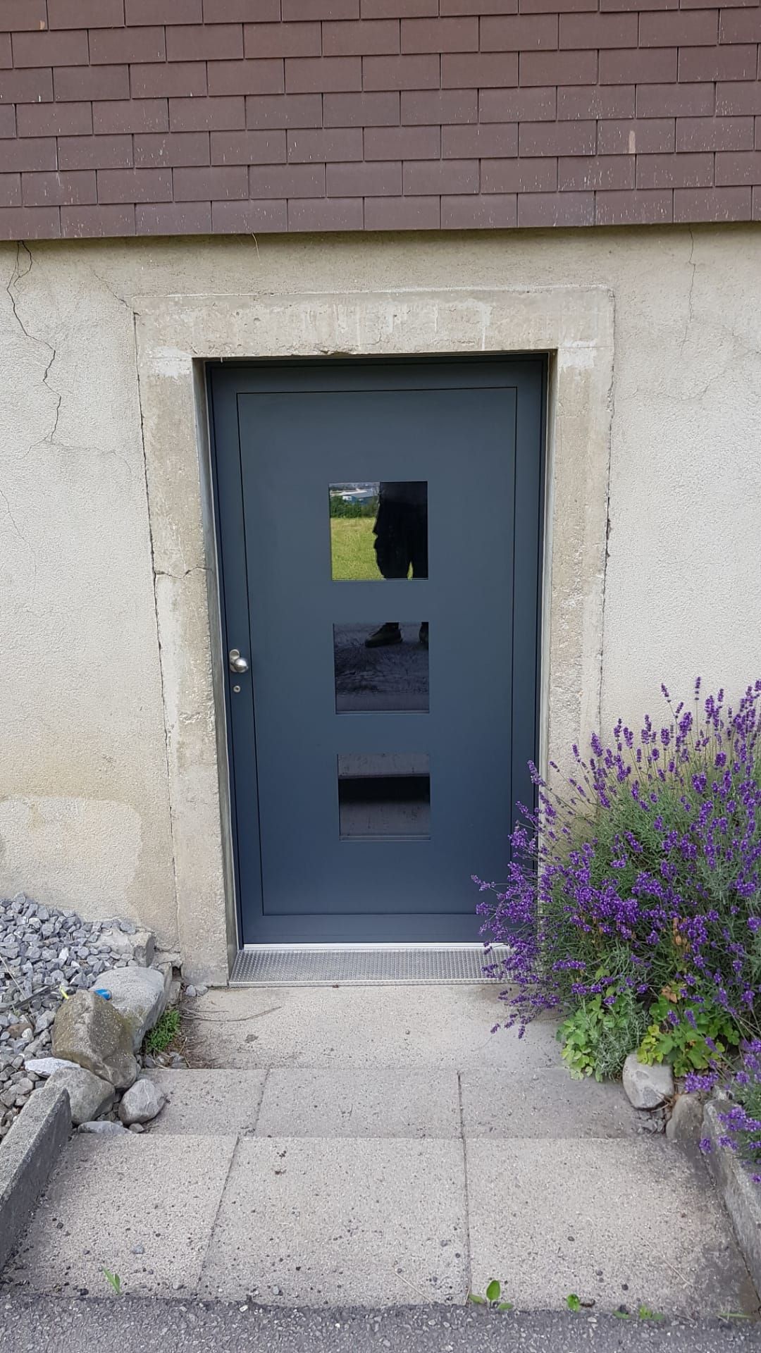 Blaugraue Haustür mit kleinen Fenstern in einer Steinmauer, umrahmt von lila Blumen und einem gepflasterten Weg