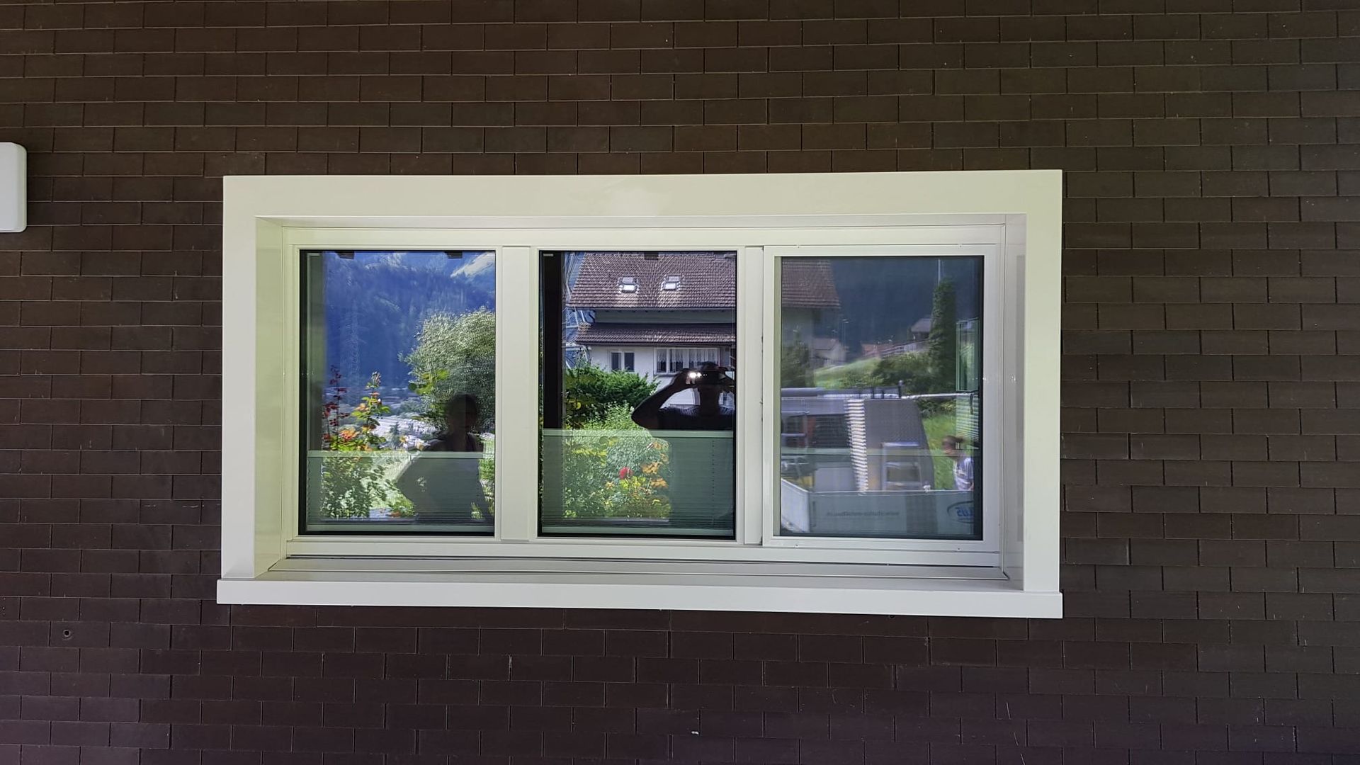 Ein dreiteiliges Fenster mit weissem Rahmen in einer Backsteinmauer spiegelt Bäume, Himmel und nahegelegene Gebäude wider.