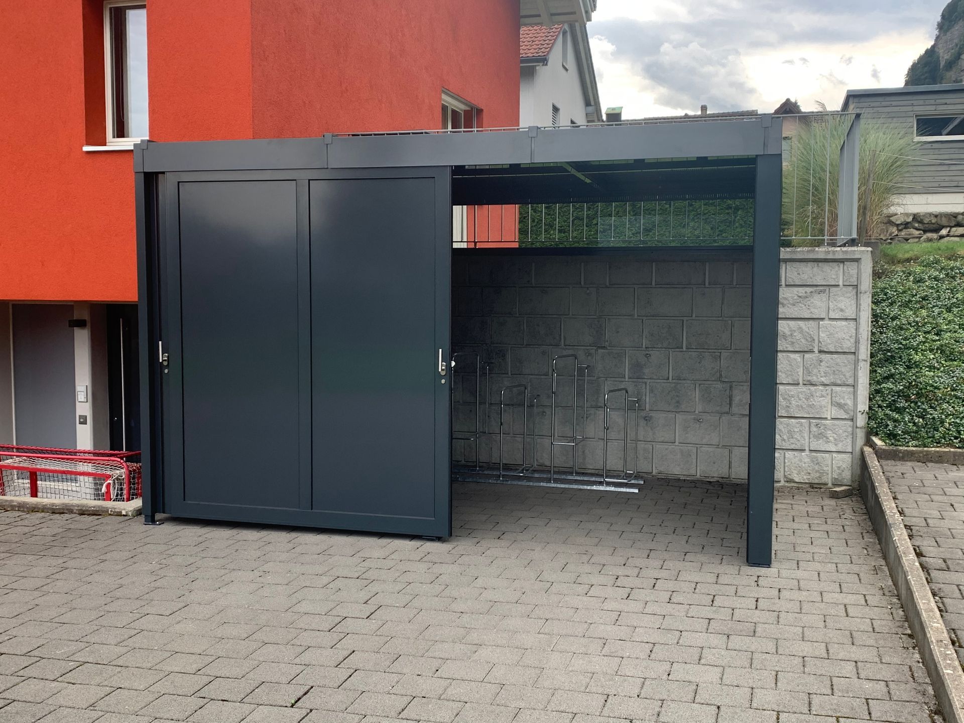 Schwarzer Fahrradunterstand aus Metall mit Schiebetür neben einem roten Gebäude und einem gepflasterten Innenhof