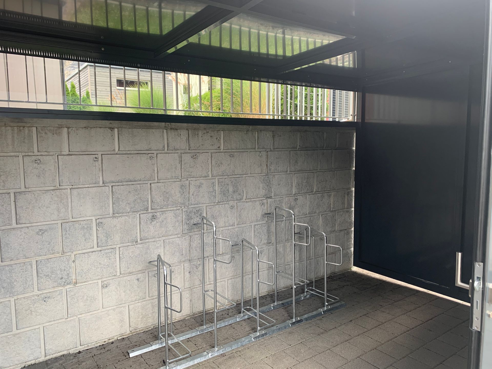 Überdachter Fahrradabstellplatz mit Metallständern vor einer grauen Betonsteinmauer und einem schwarzen Tor
