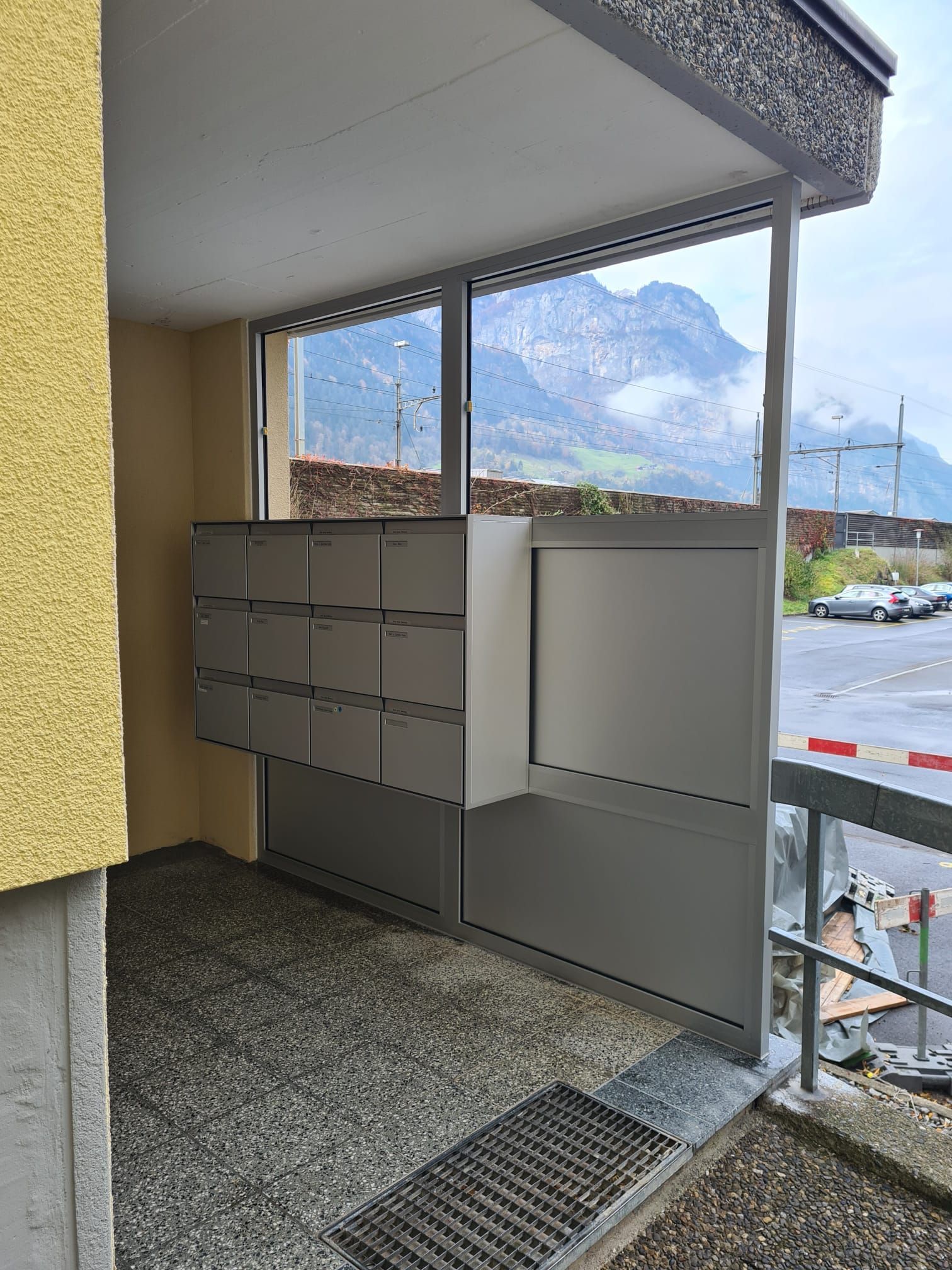 Überdachte Briefkästen im Freien neben einem Gehweg, hinter dem Gebäude sind die Berge zu sehen.
