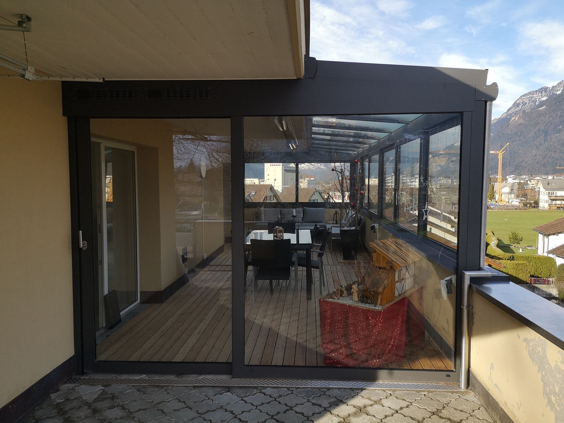 Geschlossener Balkon mit Glasfenstern, Tisch und Stühlen sowie einem roten Pflanzkübel mit Blick auf die Hügellandschaft.
