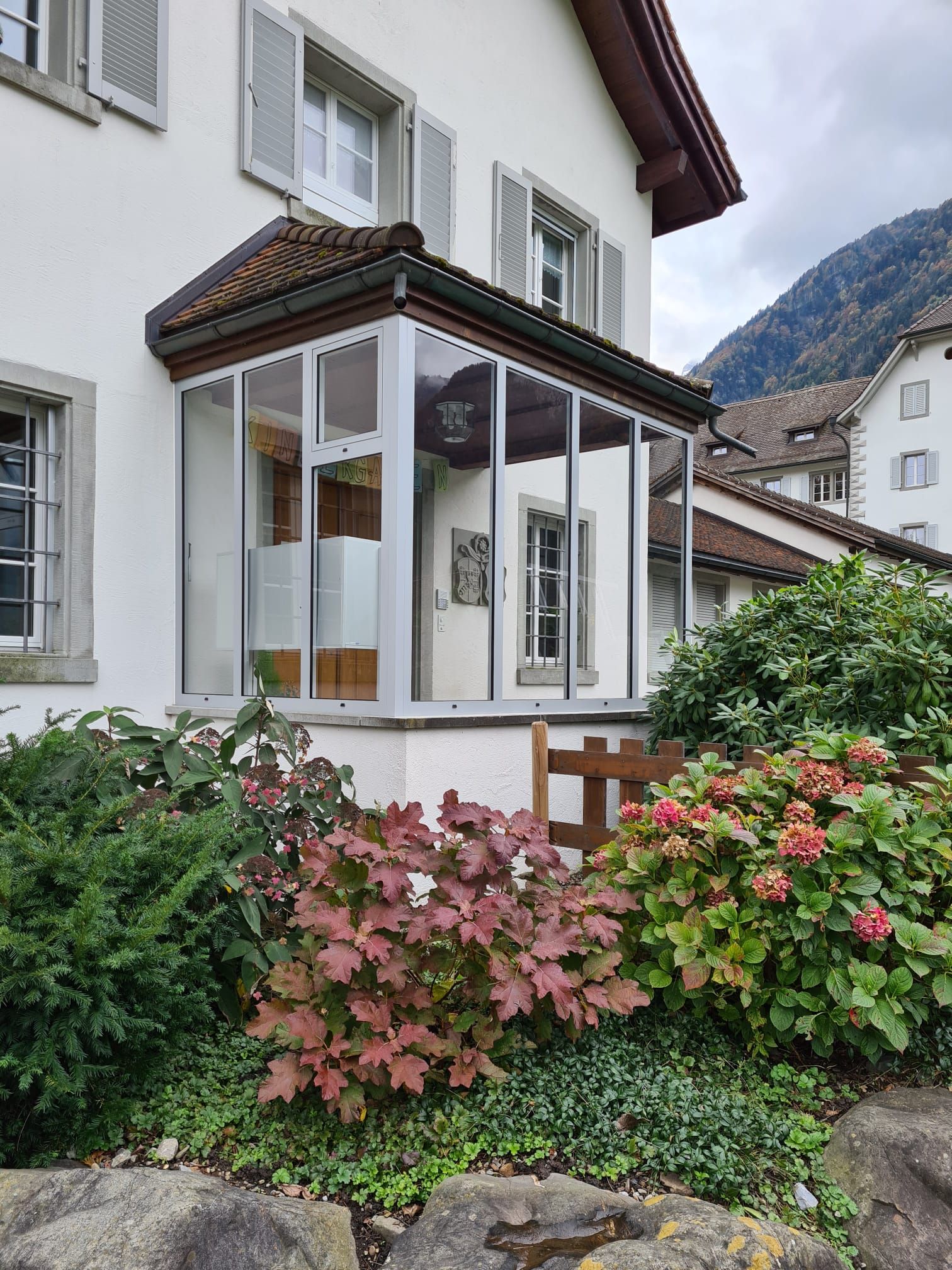 Verglaster Wintergarten an einem weissen Haus mit rosa Blumen und Bergkulisse