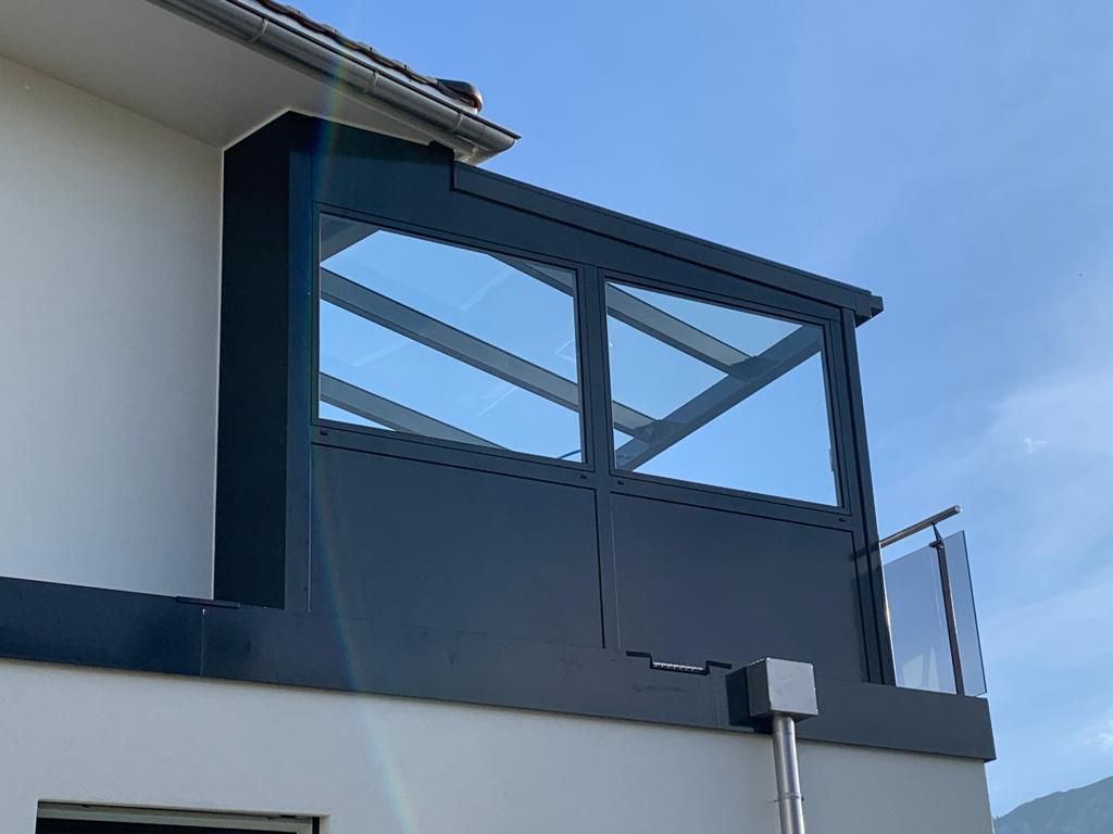 Blauer Balkon mit Glaspaneelen an einem weissen Gebäude vor klarem Himmel