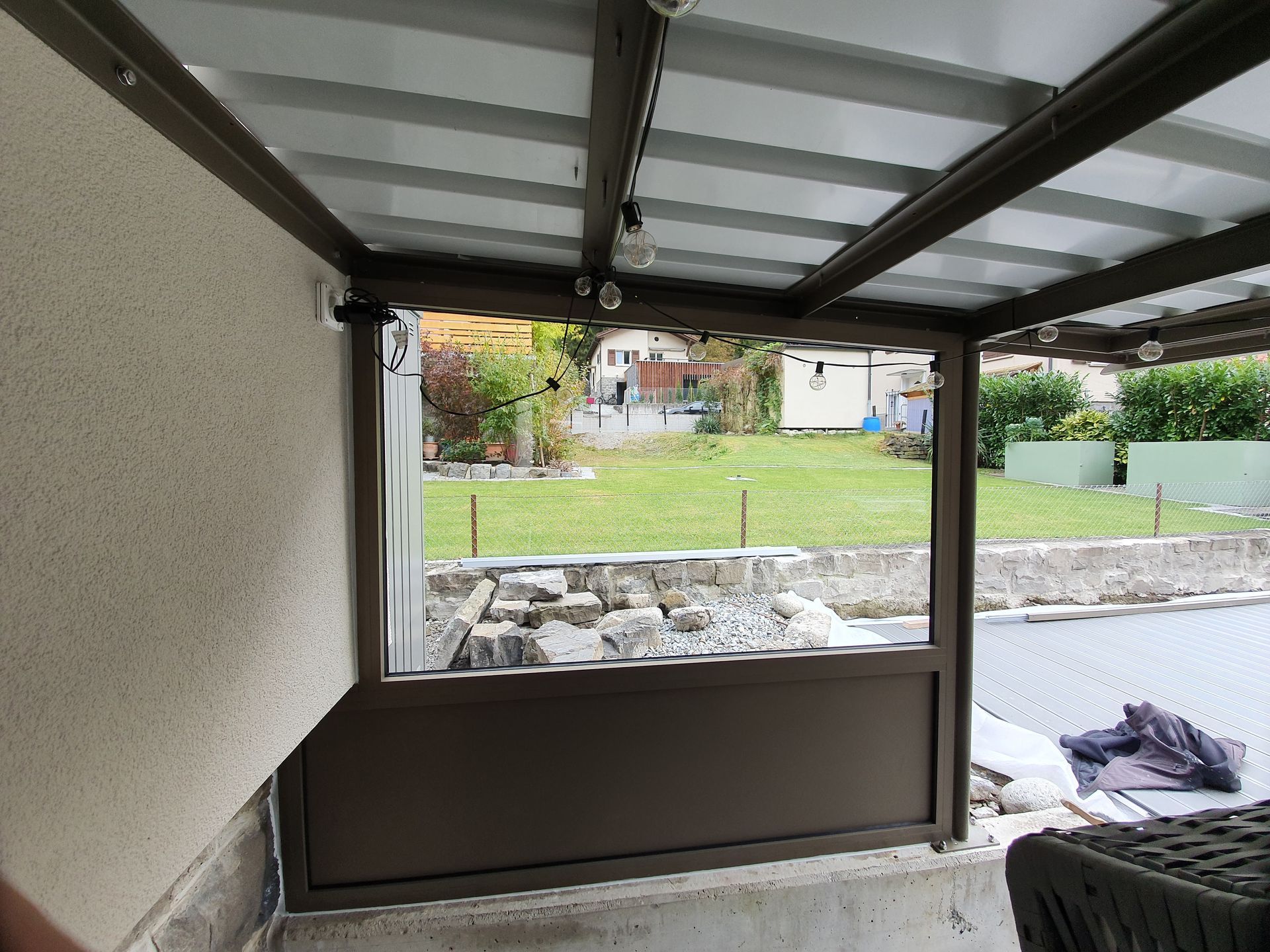 Überdachte Terrasse mit Blick auf einen Rasenhof und Häuser, mit niedriger Steinmauer und braunem Geländer.