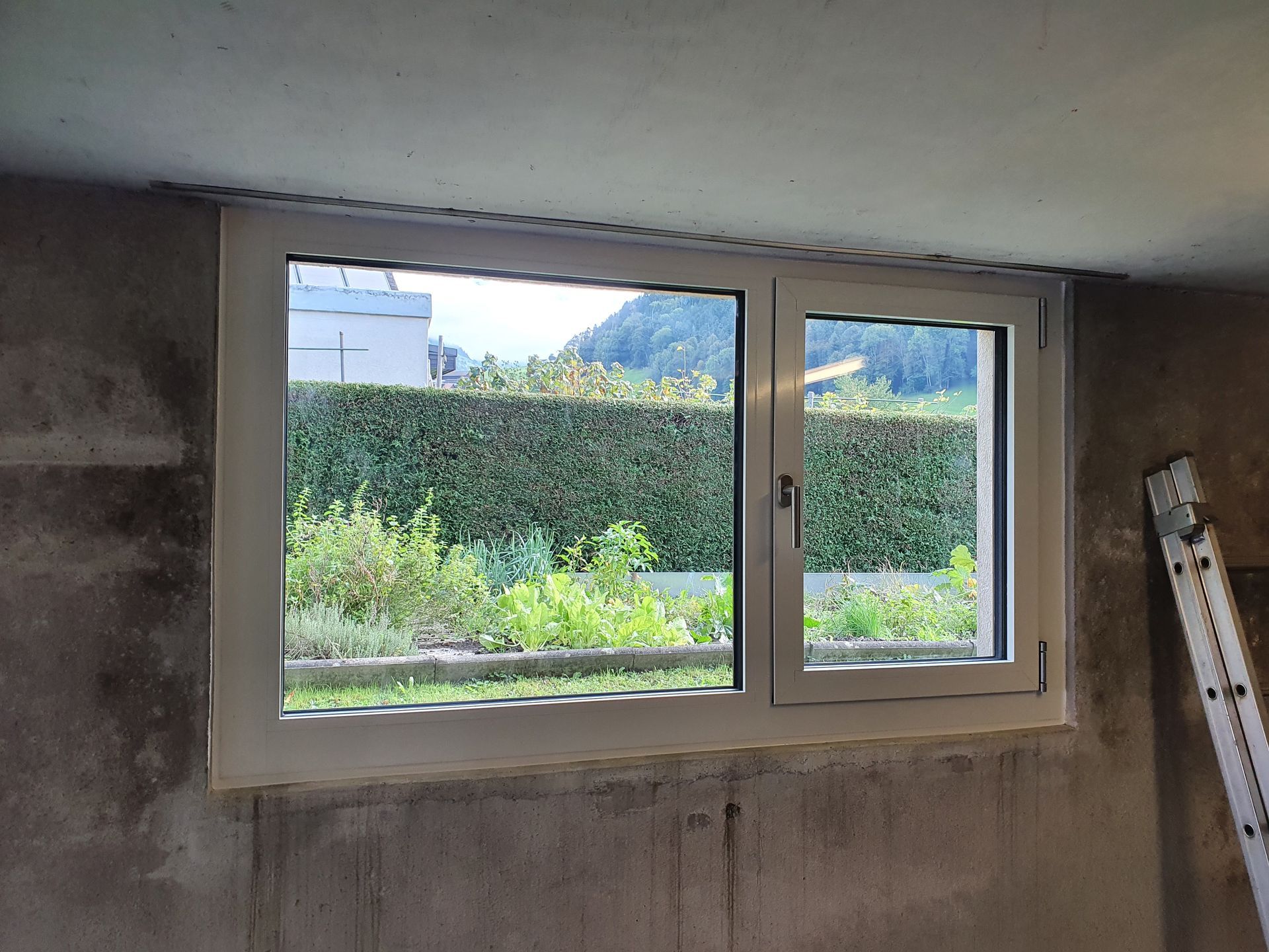 Unfertiges Zimmer mit weissem Fensterrahmen und Blick ins Grüne und auf die Hügel, rechts eine Leiter.