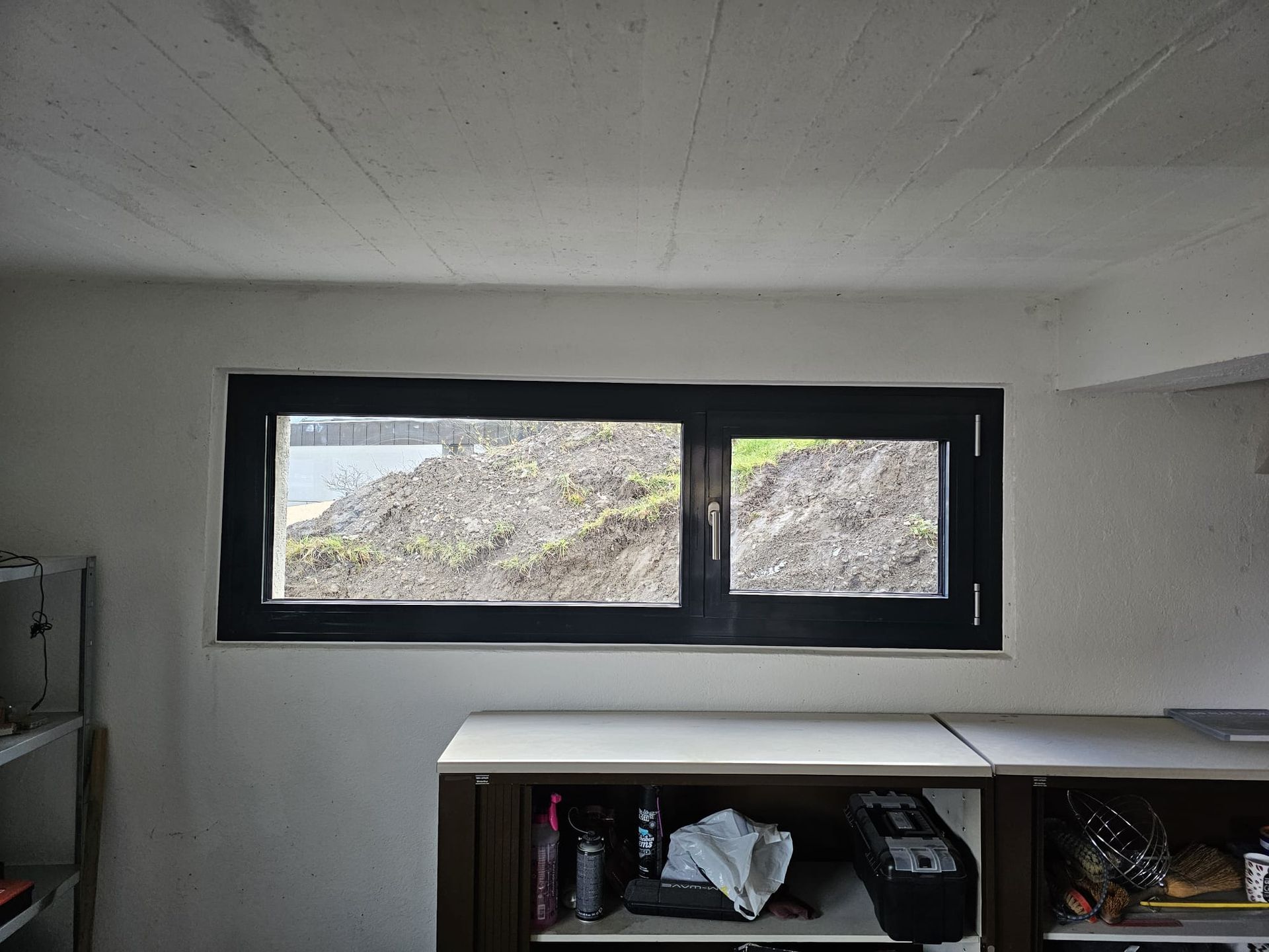 Innenraum mit einem schwarz gerahmten Fenster, weissen Wänden und einem Regal darunter.