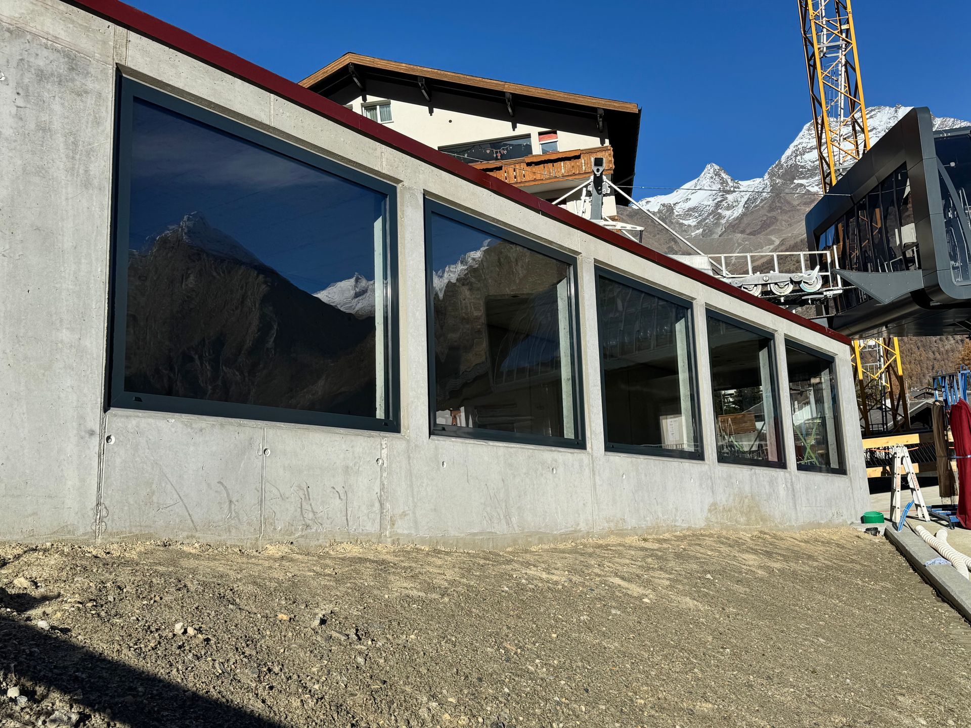 Modernes Gebäude mit grossen Fenstern an einem Felshang, im Hintergrund Berge und Baumaschinen.
