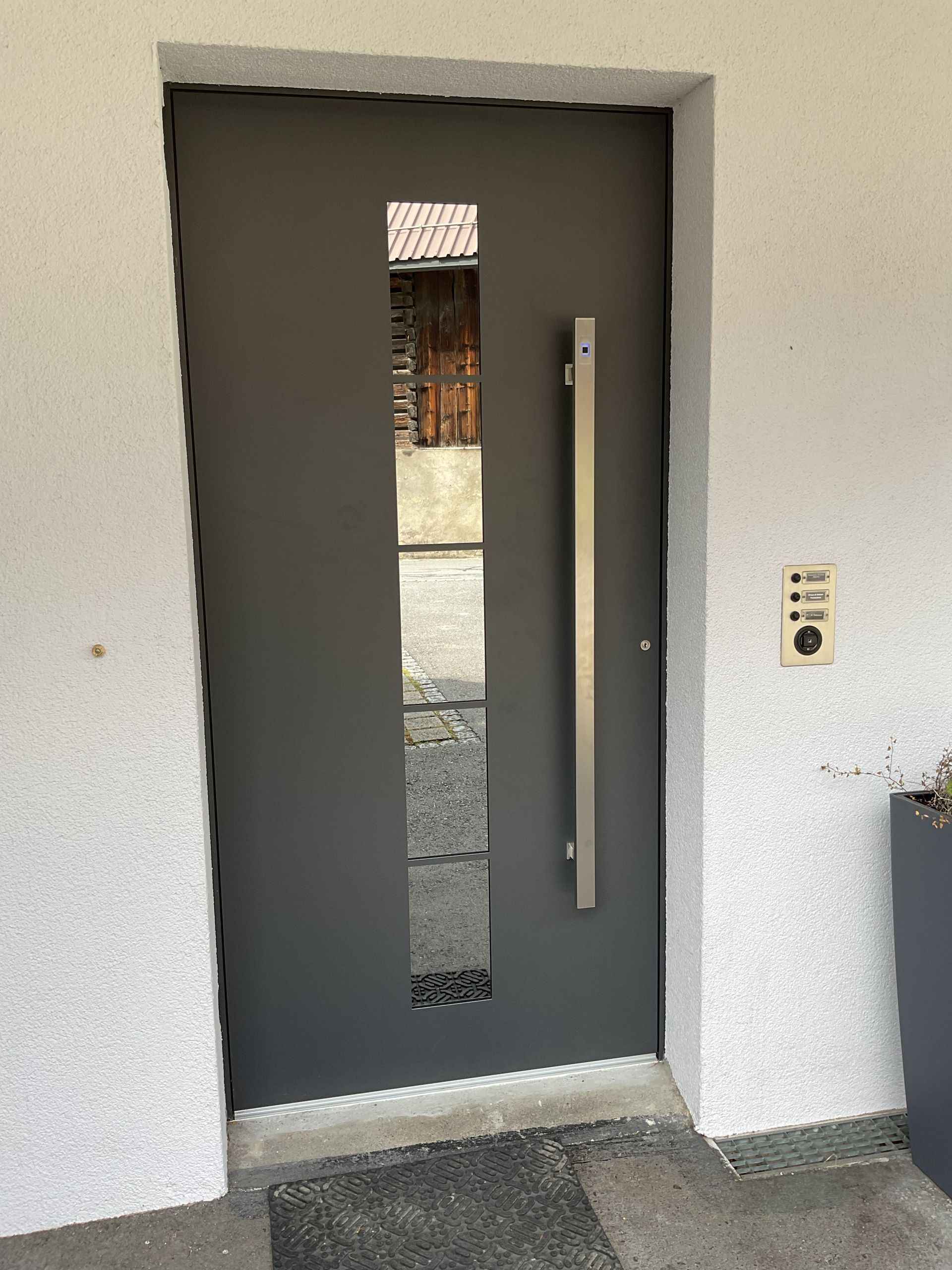 Graue Haustür mit schmaler, vertikaler Glasscheibe und an der Wand montierter Türklingel