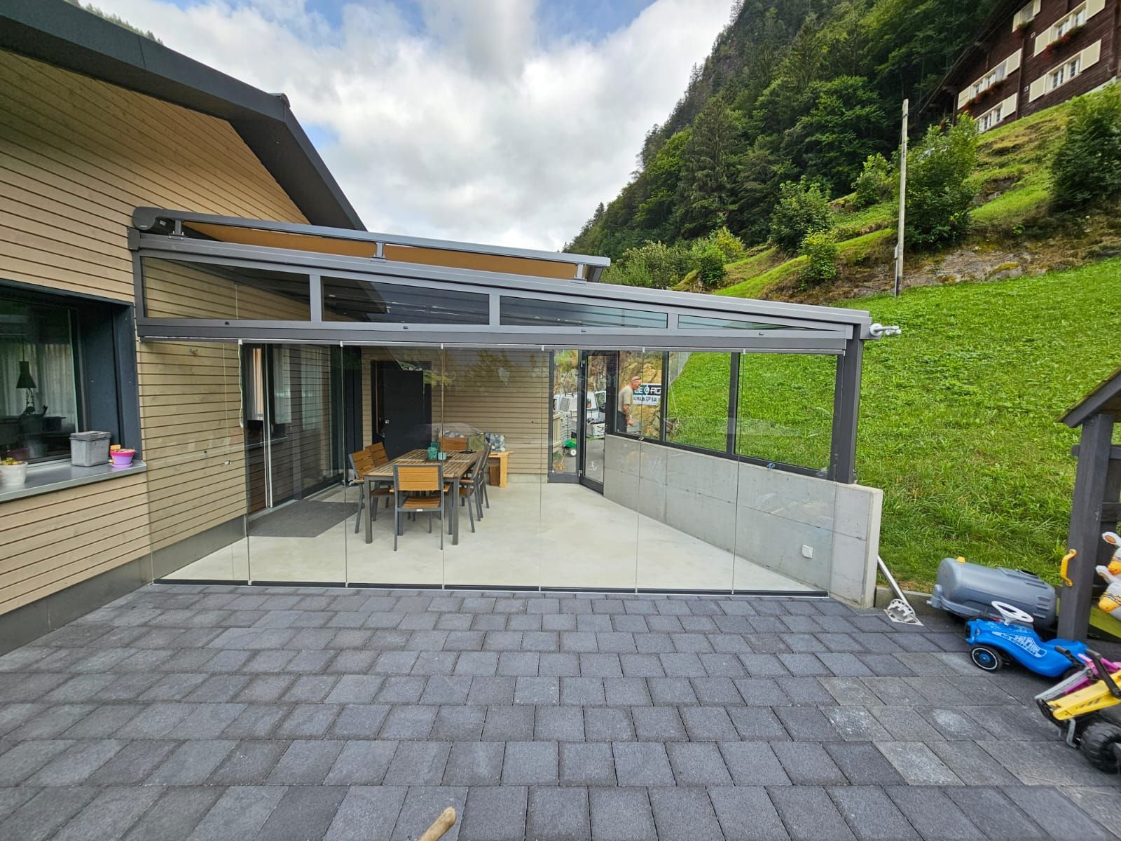 Überdachte Terrasse mit Tisch und Stühlen neben einem Haus, mit Blick auf eine gepflasterte Auffahrt und einen grünen Hang.
