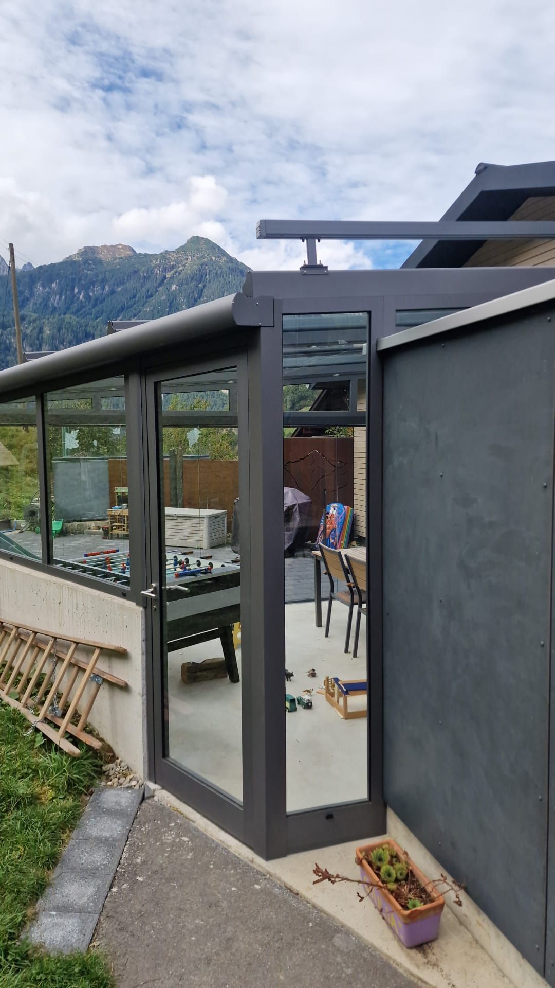 Moderne Terrasse neben einem grauen Haus, mit Glastüren, Gartenmöbeln und Bergen im Hintergrund