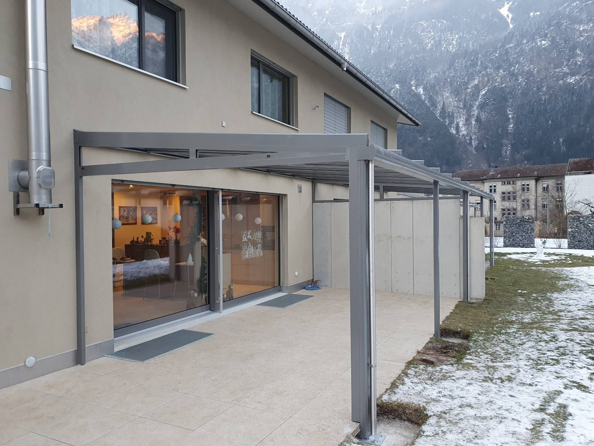 Überdachte Terrasse neben einem beigefarbenen Haus mit Glasschiebetüren und Blick auf eine schneebedeckte Berglandschaft.