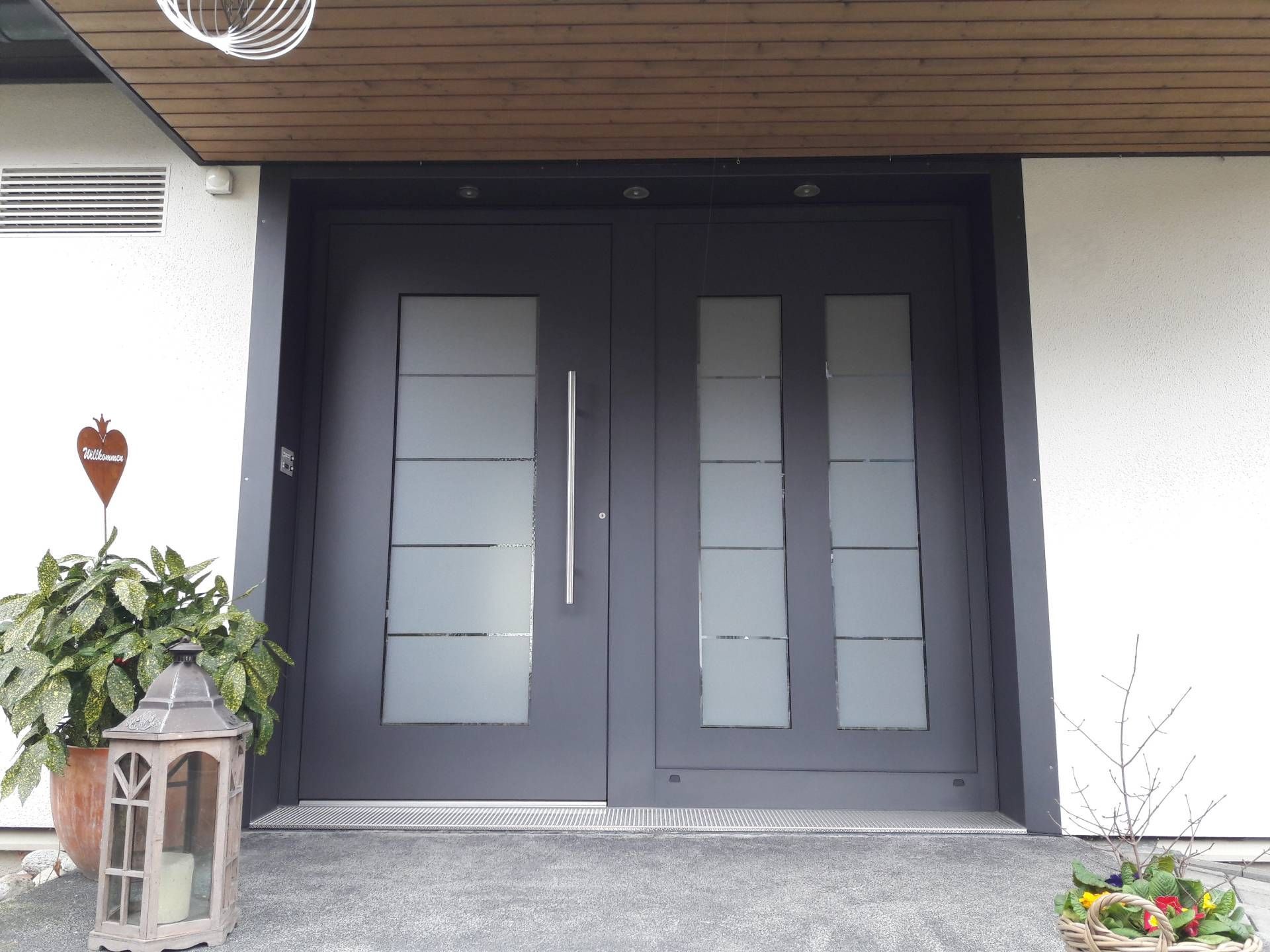 Moderne, dunkle Haustür mit Milchglasscheiben unter einem hölzernen Vordach, flankiert von Topfpflanzen