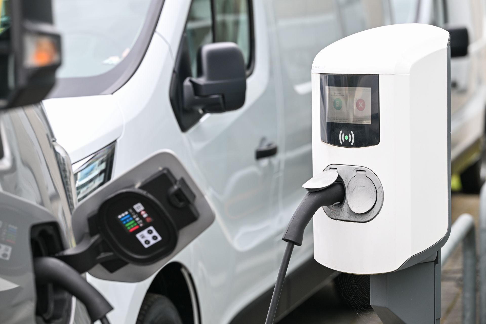 Fourgonnette blanche en charge à une borne de recharge pour véhicules électriques ; câble gris branché ; environnement extérieur.