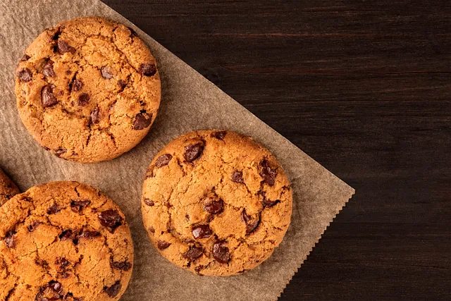 Tres galletas con chispas de chocolate están colocadas sobre un trozo de papel encerado sobre una mesa de madera.