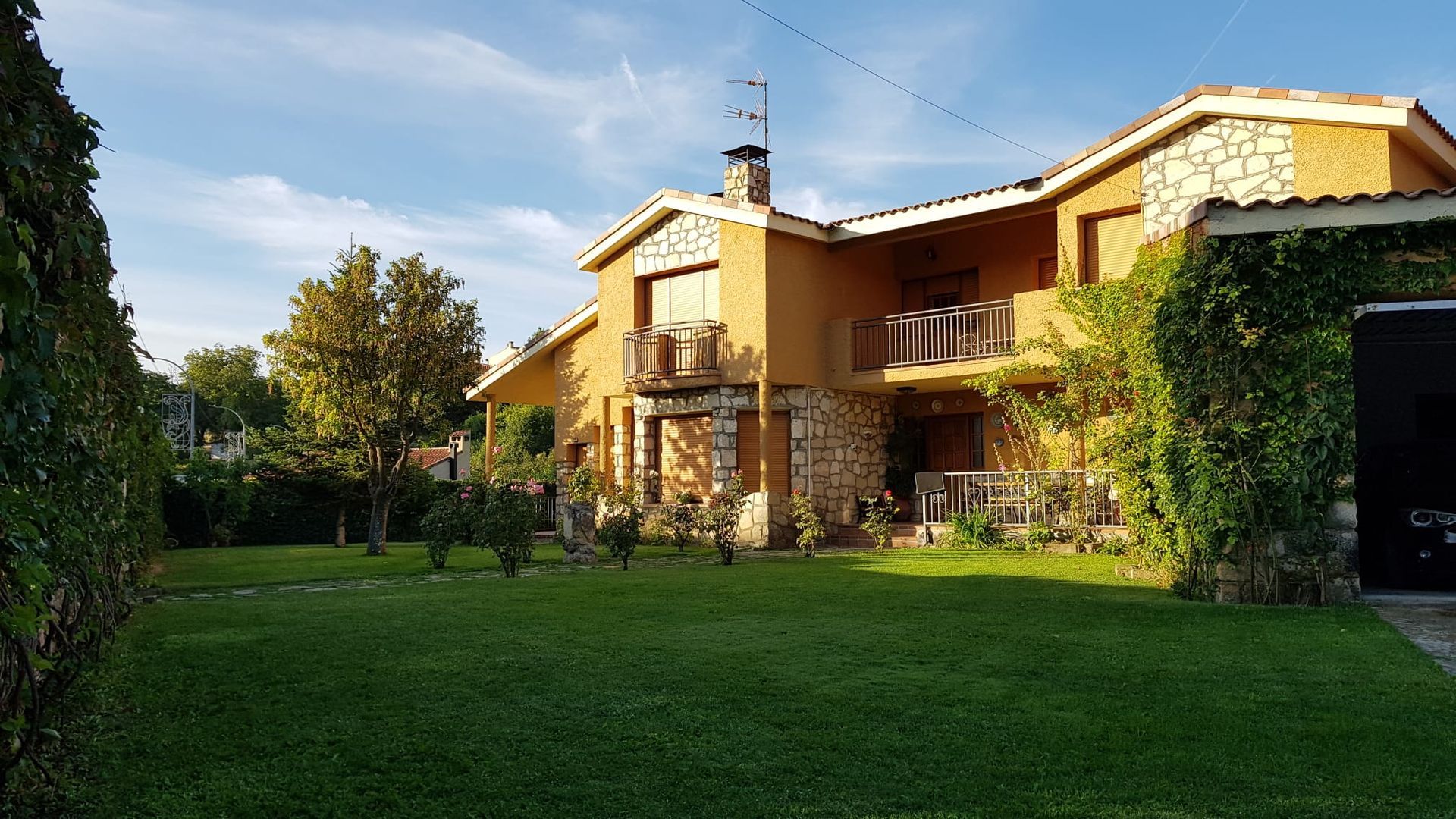 Casa de dos pisos con paredes color canela, detalles de piedra y balcones, ubicada en un patio verde bajo un cielo azul.