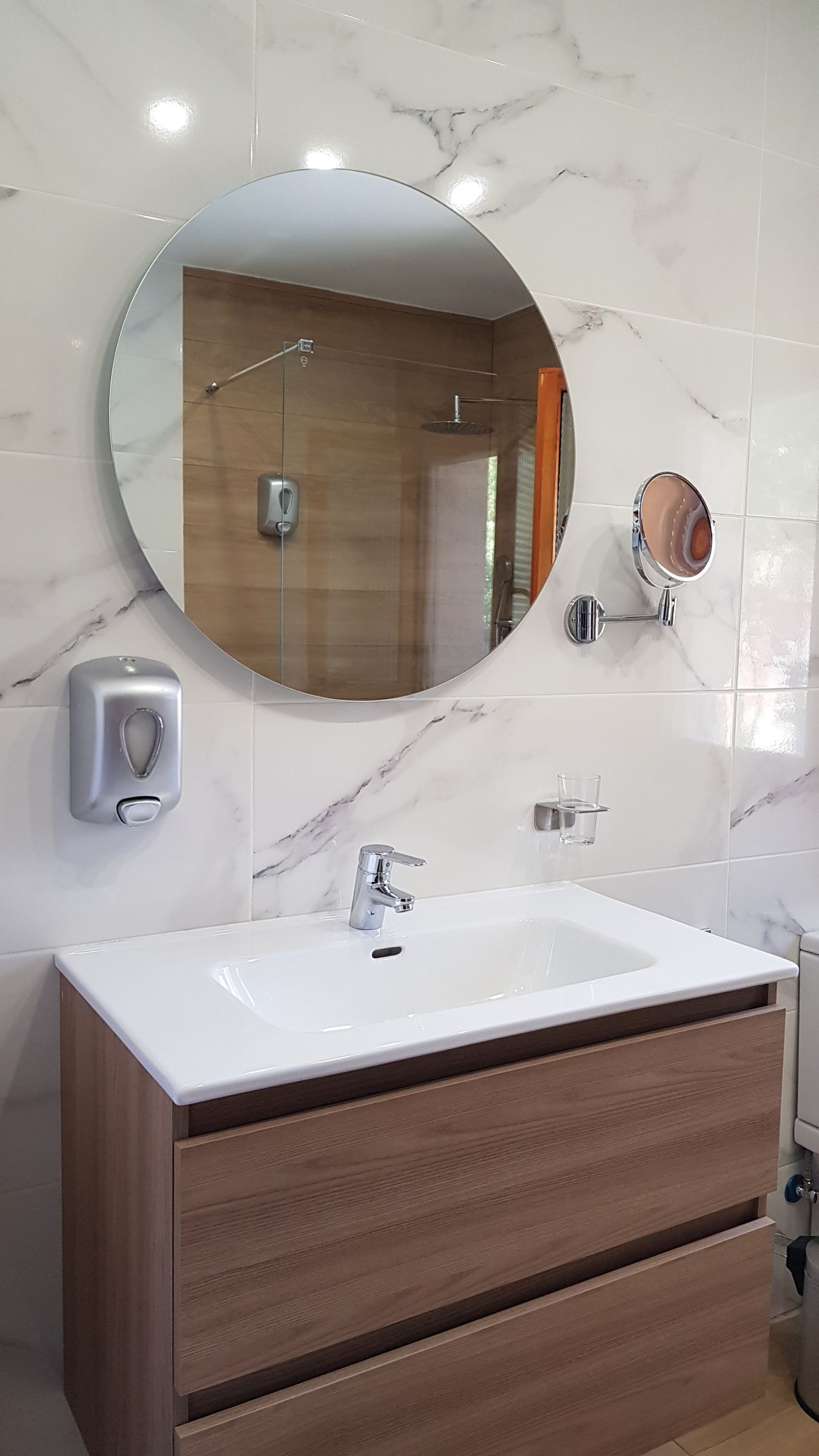 Mueble de baño con espejo redondo, lavabo y dispensador de jabón. Azulejos imitación mármol blanco y mueble de madera.