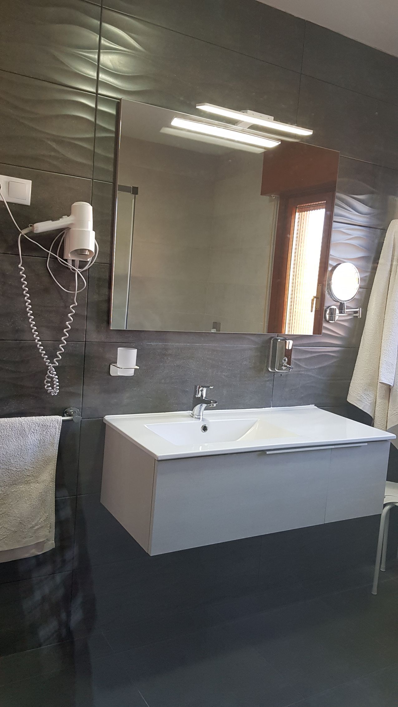 Baño con lavabo blanco, espejo grande y pared con textura oscura. Un secador de pelo colgado a la izquierda.