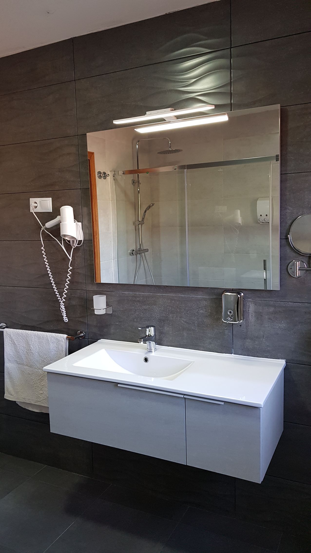 Baño con lavabo blanco, espejo grande y paredes de azulejos grises. Incluye ducha y secador de pelo.