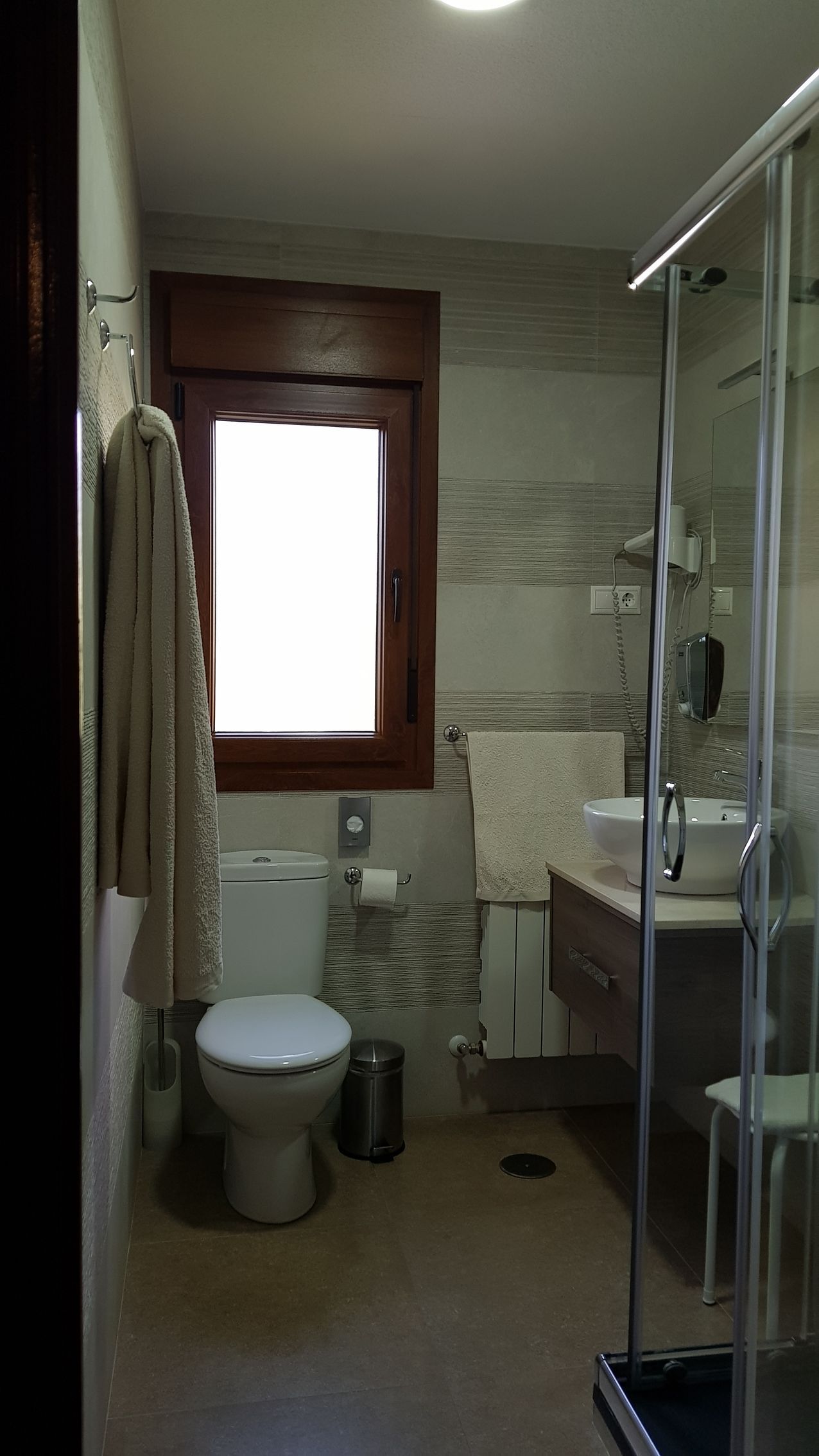 Baño con inodoro, lavabo, ducha y ventana. Tonos beige y marrón.